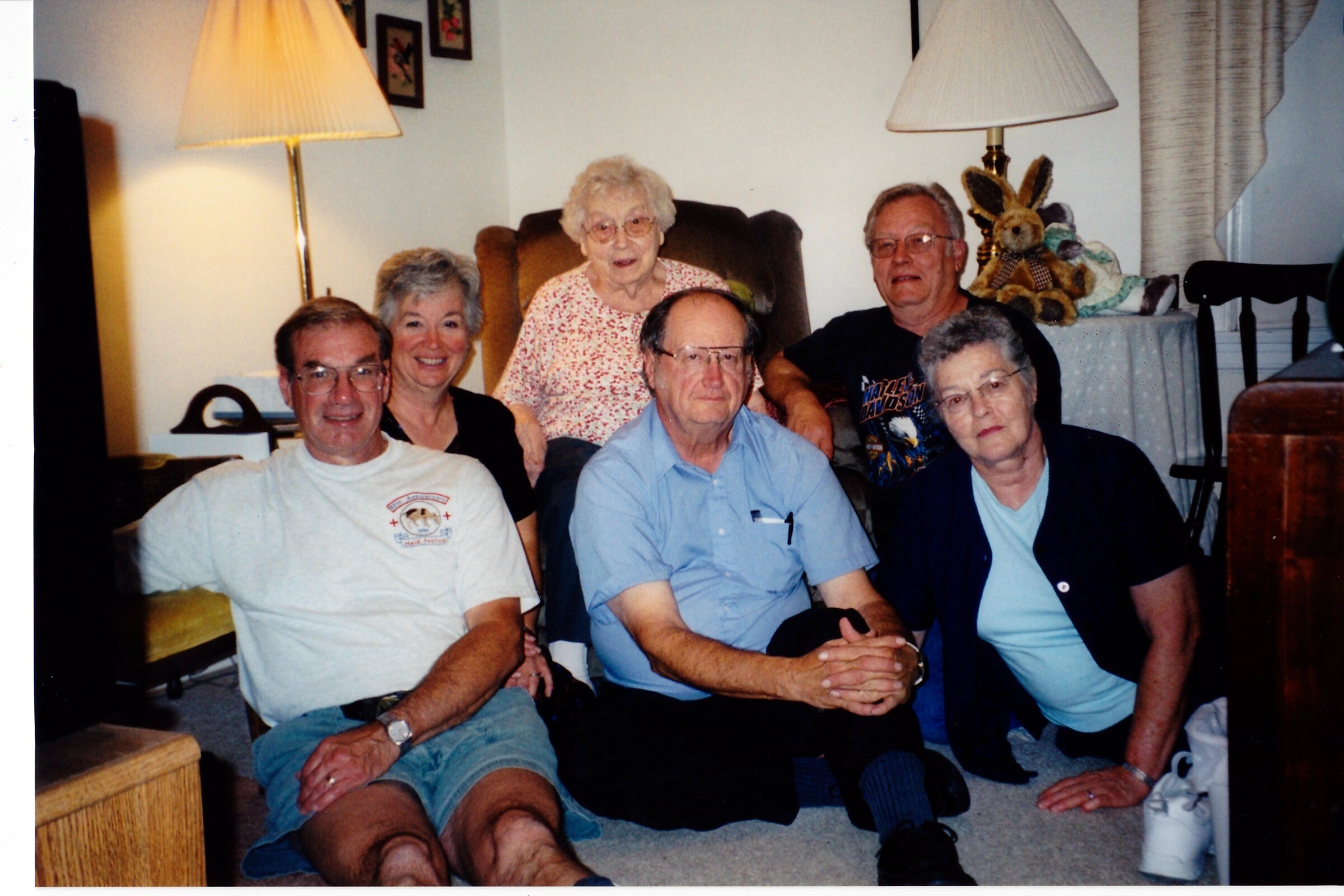 Six adults gathered in a living room posing for a group photo, smiling at the camera.
