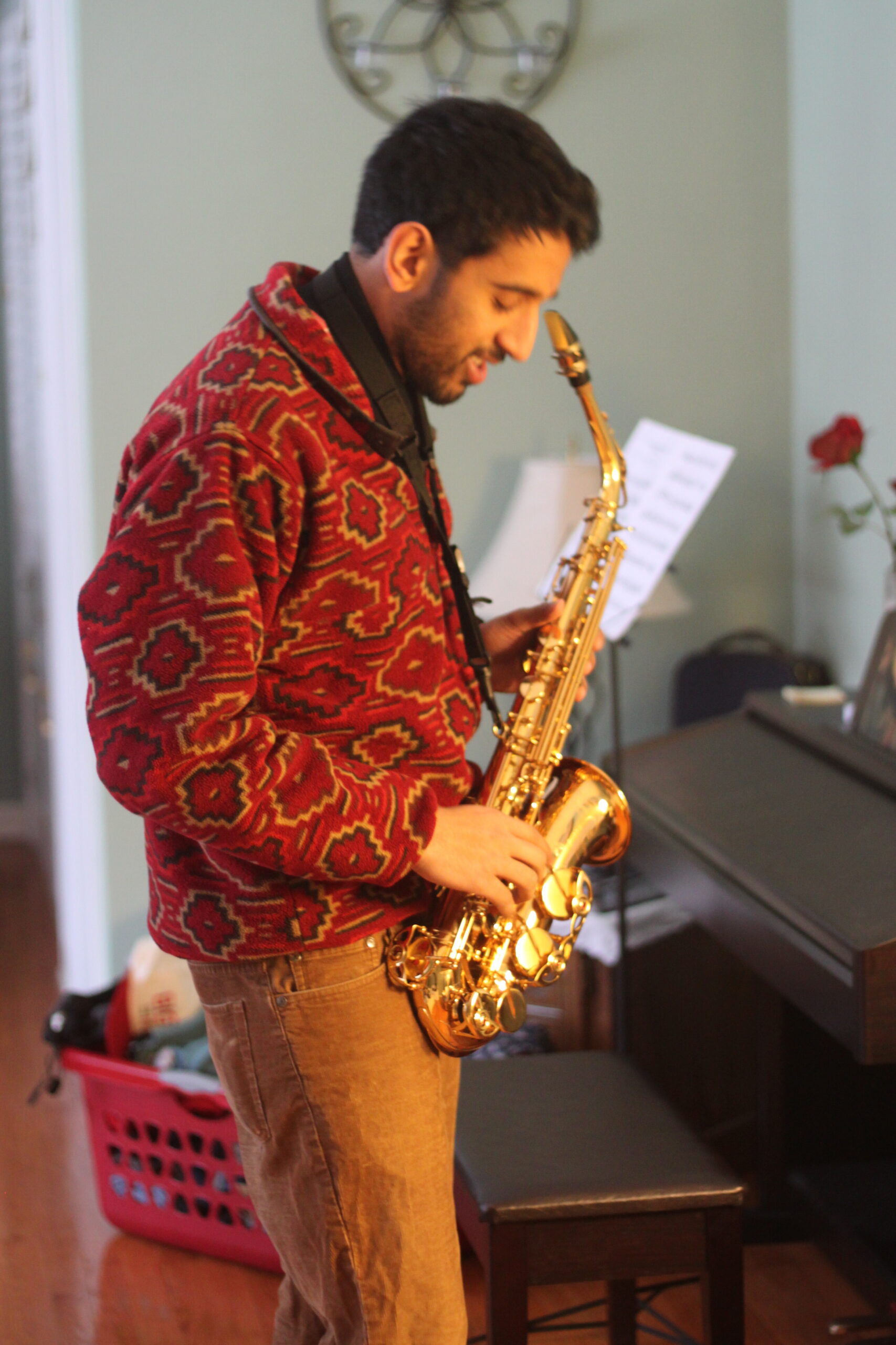 Man in a red patterned jacket playing a gold saxophone indoors, looking down at the instrument and sheet music nearby.