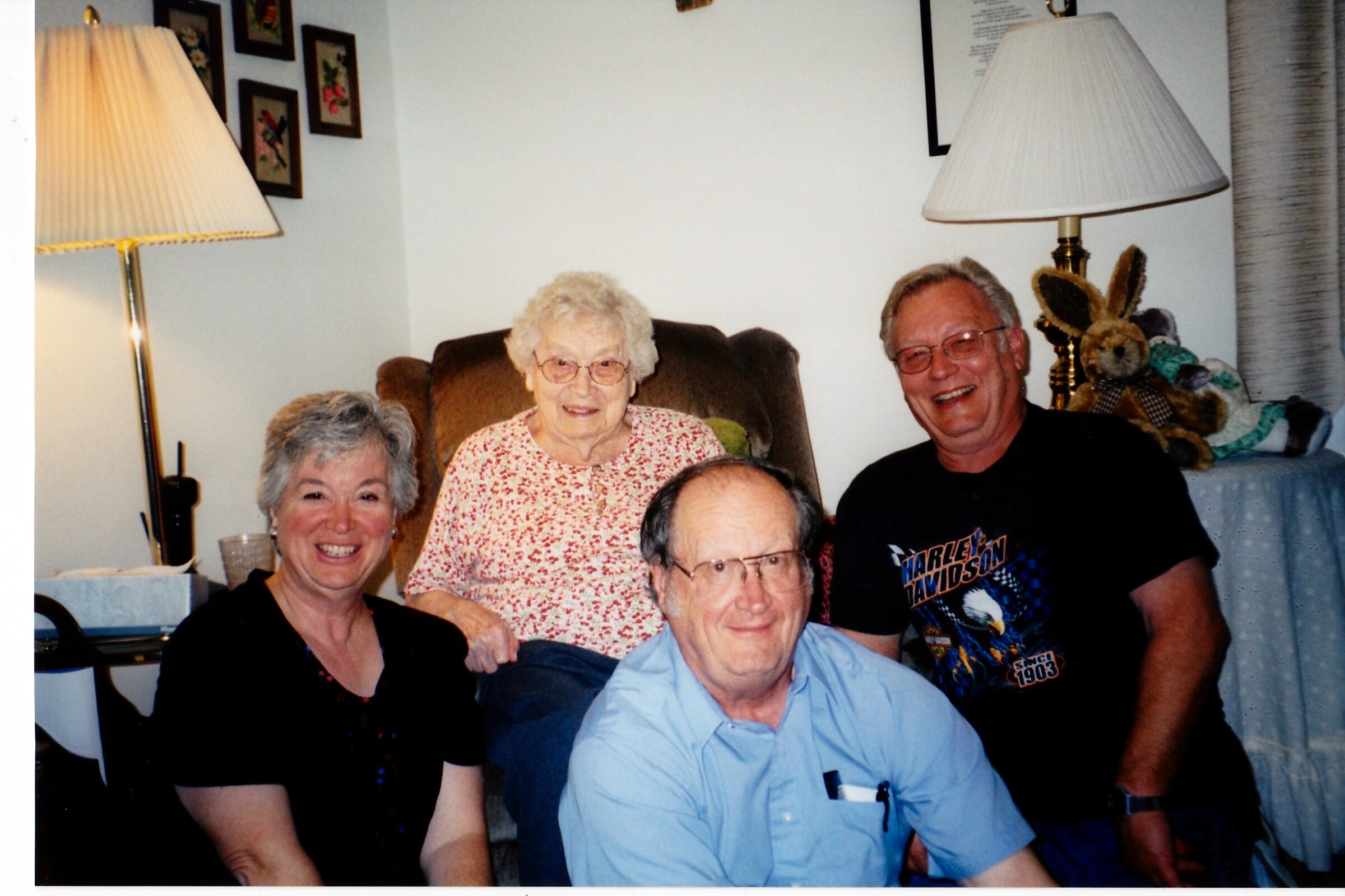 Smiling group of five adults posing for a casual family photo in a living room, with lamps and a teddy bear in the background.