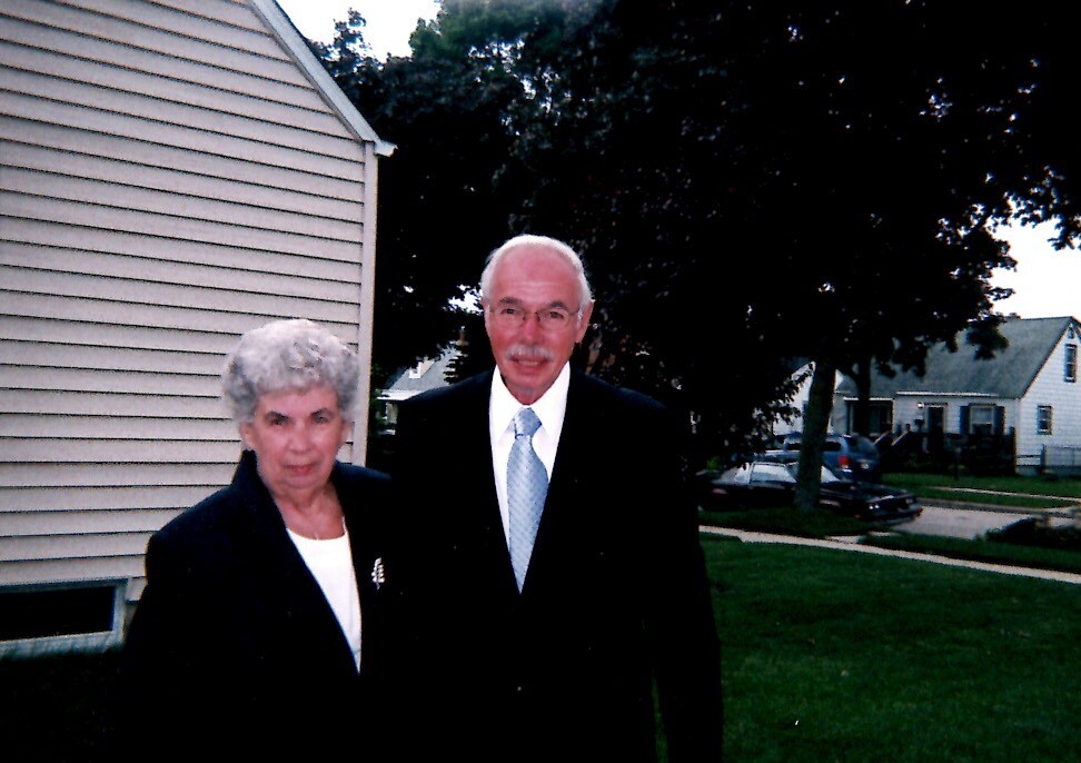 Smiling elderly man in a dark suit and light blue tie beside an elderly woman in a black blazer outside a beige house in a suburban yard.