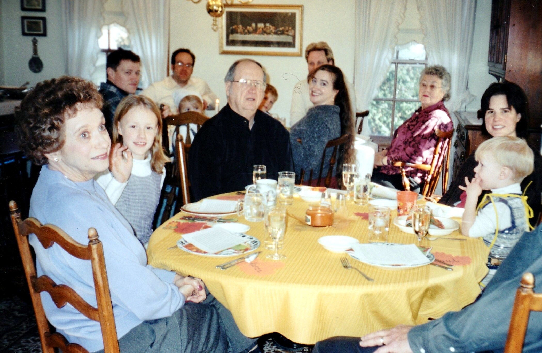 Group of people around a round dining table in a home, smiling during a family meal with plates and glasses visible in the scene.