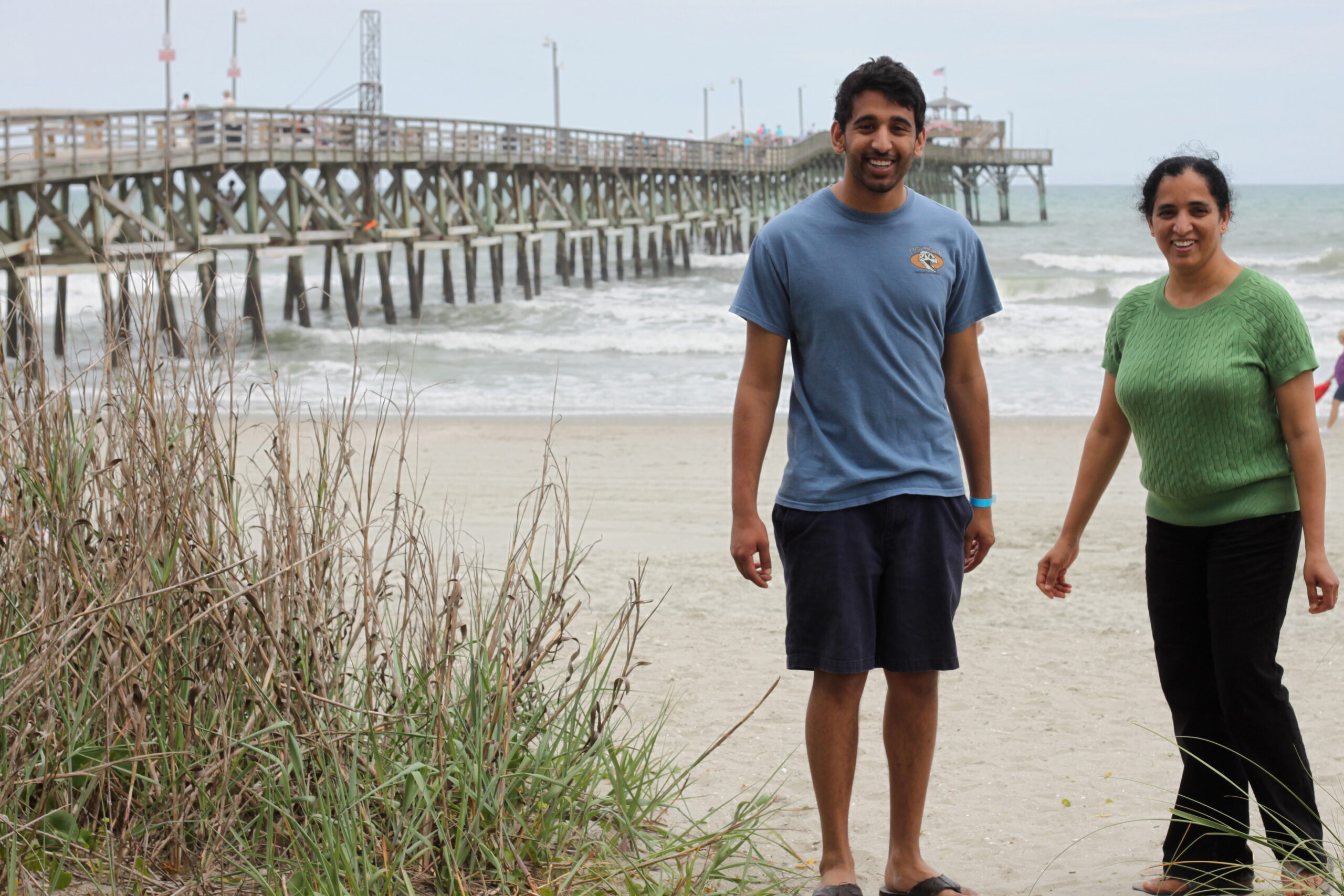Two smiling people stand on a sandy beach with a long wooden pier extending into the ocean behind them, and dune grass in the foreground.