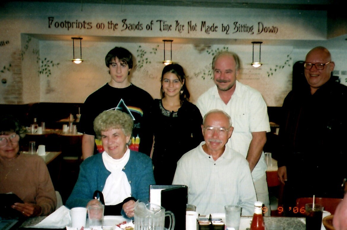 Six people around a restaurant table posing for a group photo, with a decorative wall and sign in the background reading about footprints.