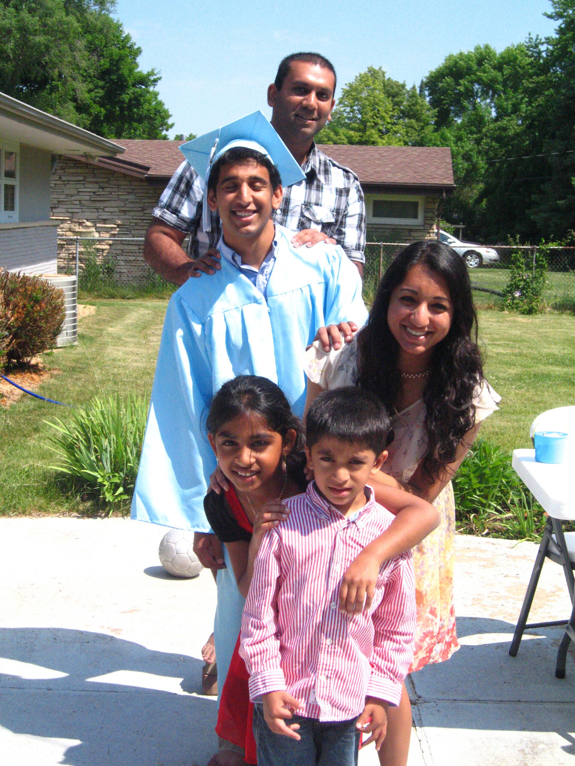 Smiling graduate in a blue cap and gown posing with a man and several children outdoors on a sunny day in a yard.