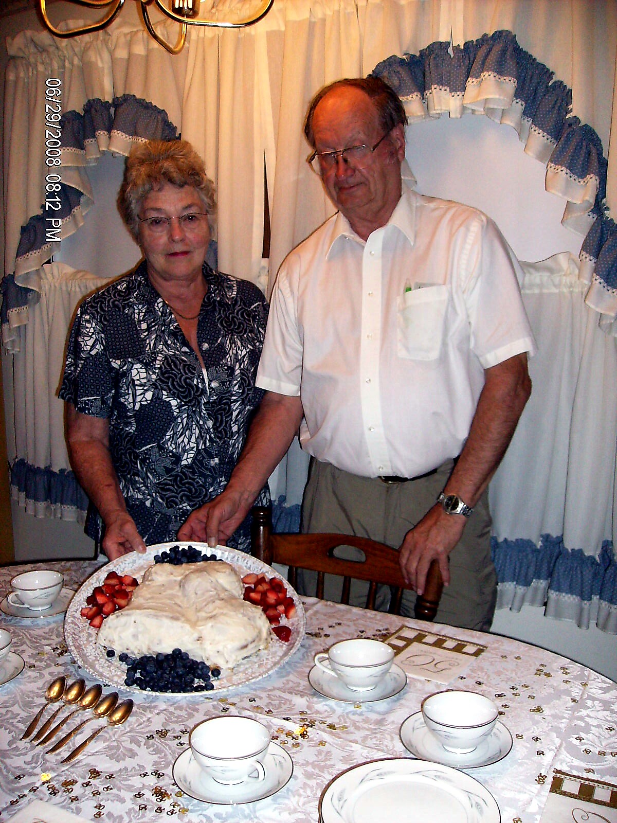 An elderly couple stands beside a cake topped with berries on a dining table during a celebration.