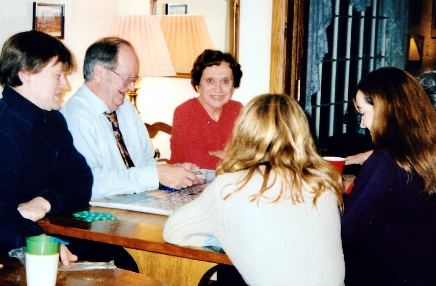 Five adults sit around a wooden dining table, playing a board game and chatting in a living room with a lamp in the background.