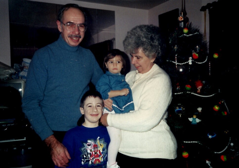 Family of four posing by a Christmas tree: grandfather in a blue turtleneck, grandmother holding a toddler, and a smiling older child at the front.