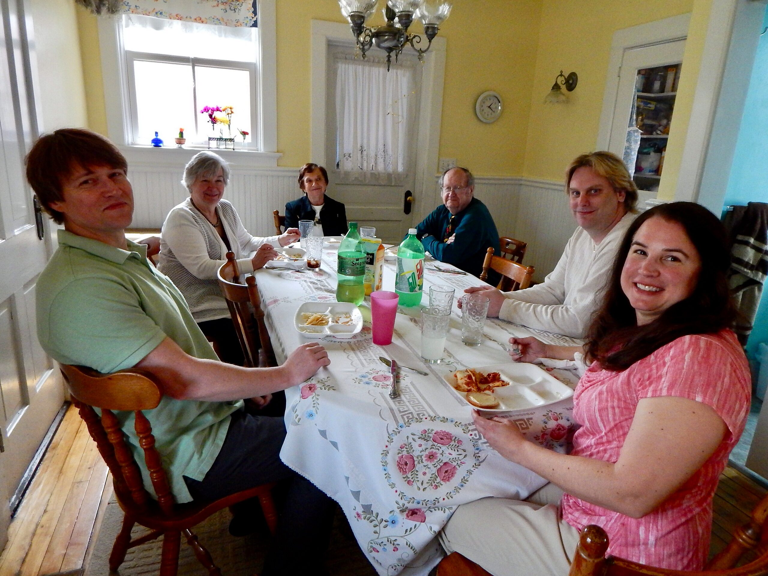 Six adults sit around a long dining table in a yellow-kitchen, sharing a meal and smiling at the camera.
