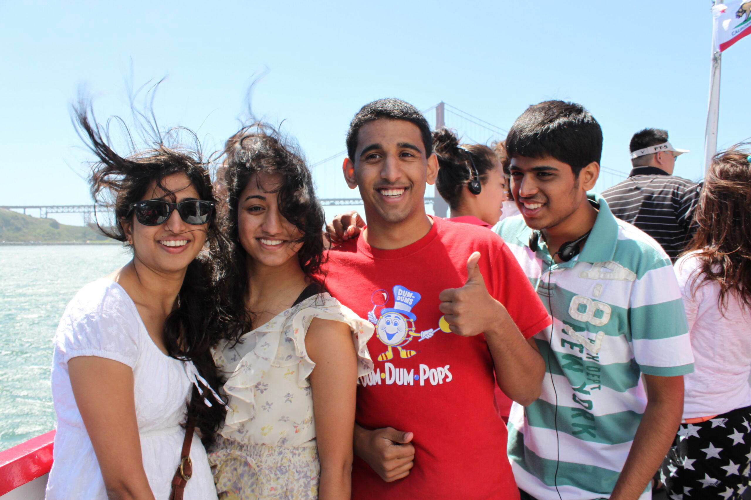 Group of friends posing for a photo on a boat with water and a bridge in the background, smiling at the camera.