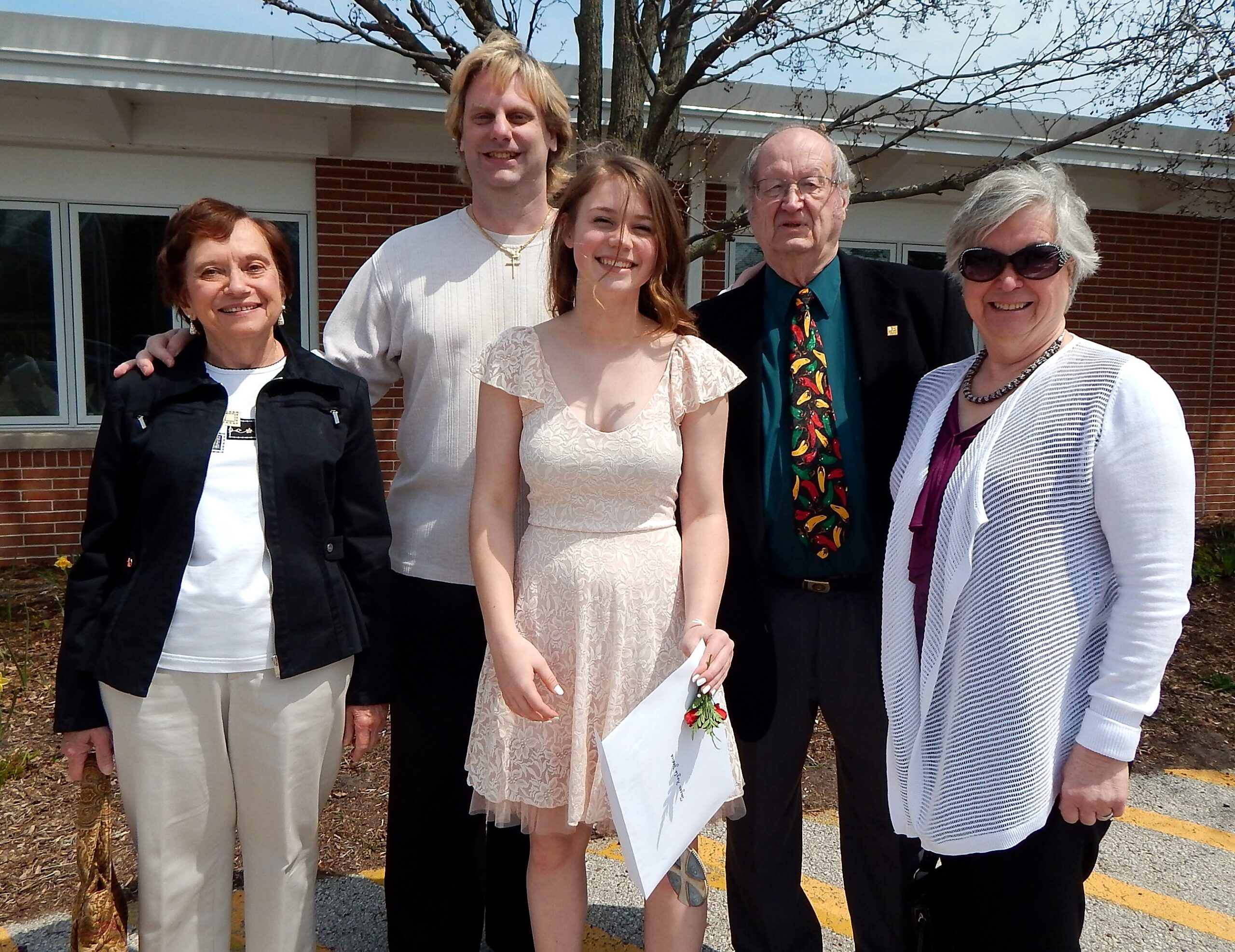 Five adults posing together outdoors in front of a brick building, smiling for the camera; from left to right: an older woman in a black jacket, a tall man in a light knit sweater, a young woman in a lace dress holding a bouquet, an older man in a dark suit with a colorful tie, and a woman with sunglasses in a white cardigan.