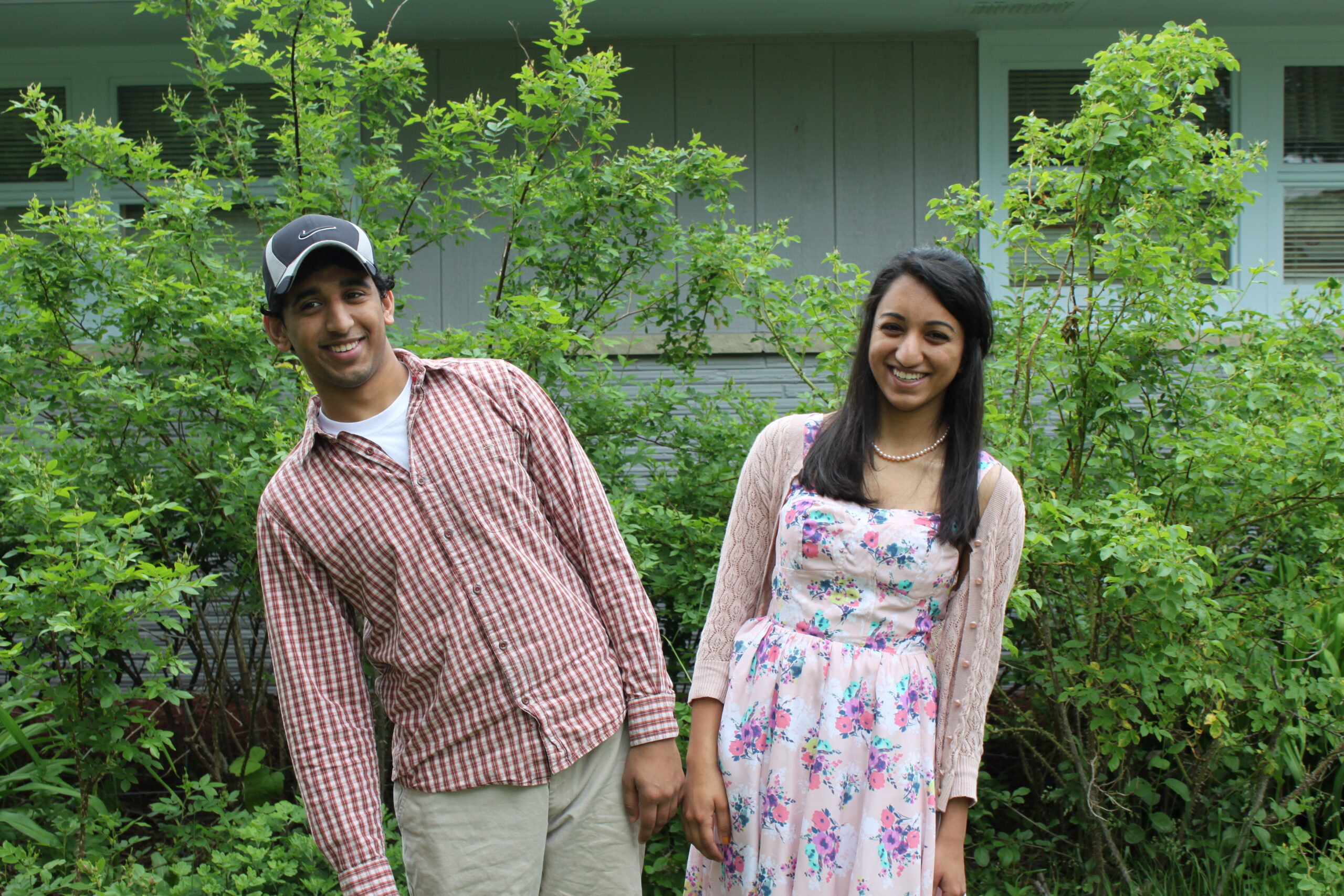 Two people stand outdoors among green bushes, smiling at the camera, holding hands lightly.