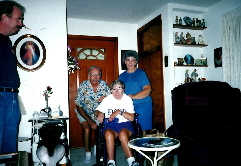 An elderly woman in a wheelchair sits center, flanked by a man in a colorful shirt and a woman in blue, in a living room pose for a photo.