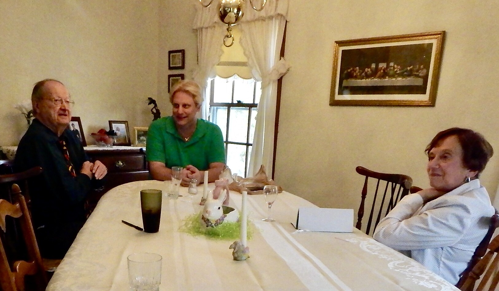 Three adults sit around a dining table in a light-colored room, with an Easter bunny centerpiece and tall candles on the table.