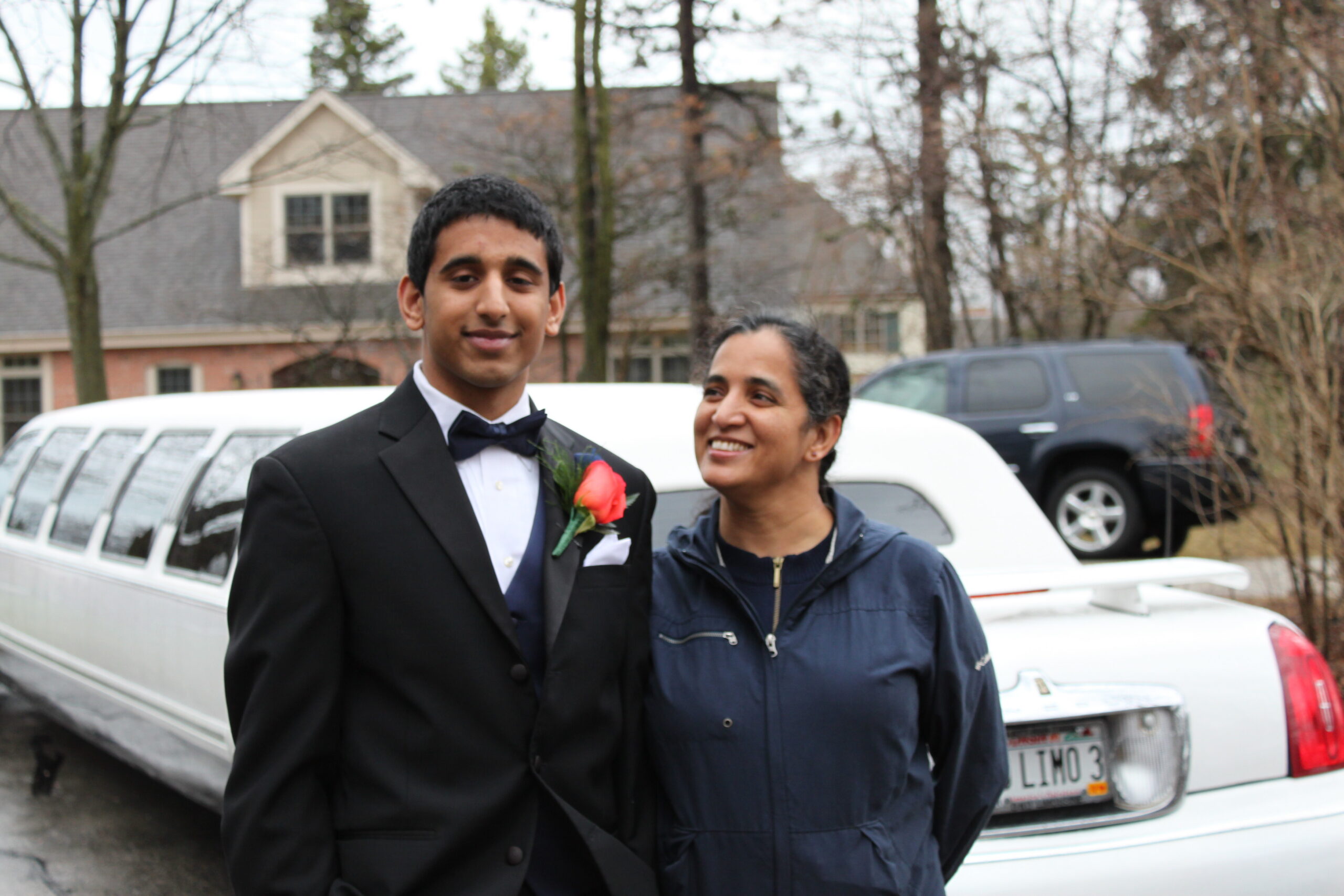 Young man in a tuxedo with a pink boutonniere poses with a smiling woman outside a white limousine in a driveway area.