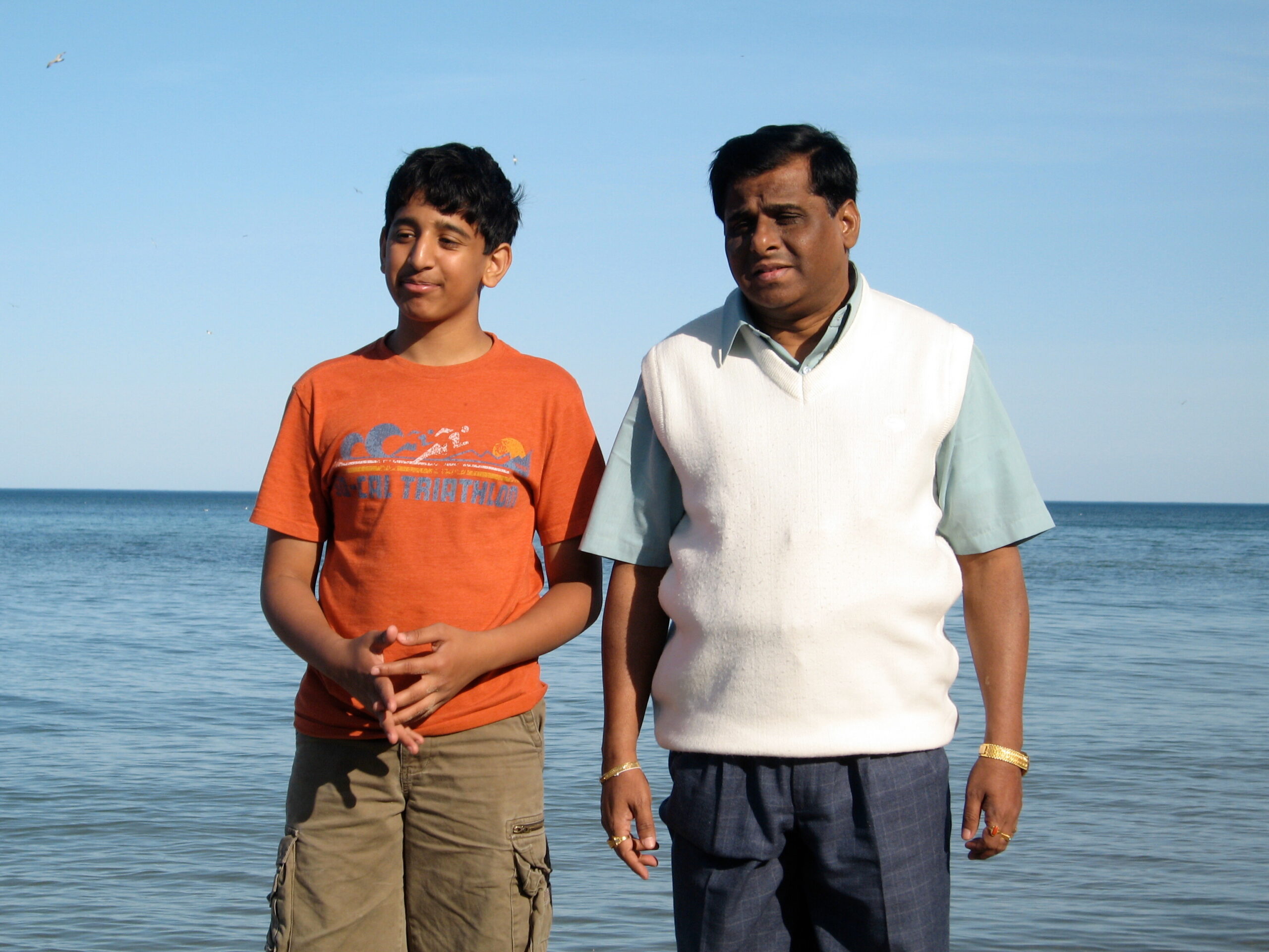 Two people stand side by side on a beach with the calm blue ocean behind them, facing slightly to the left.