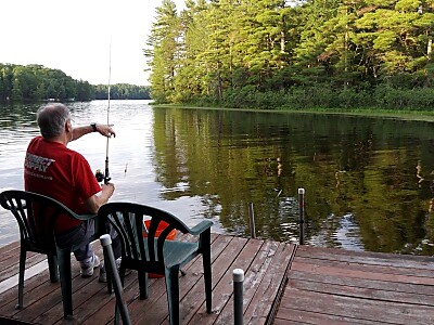 Man in a red shirt sits on a wooden dock, fishing into a calm lake bordered by trees.