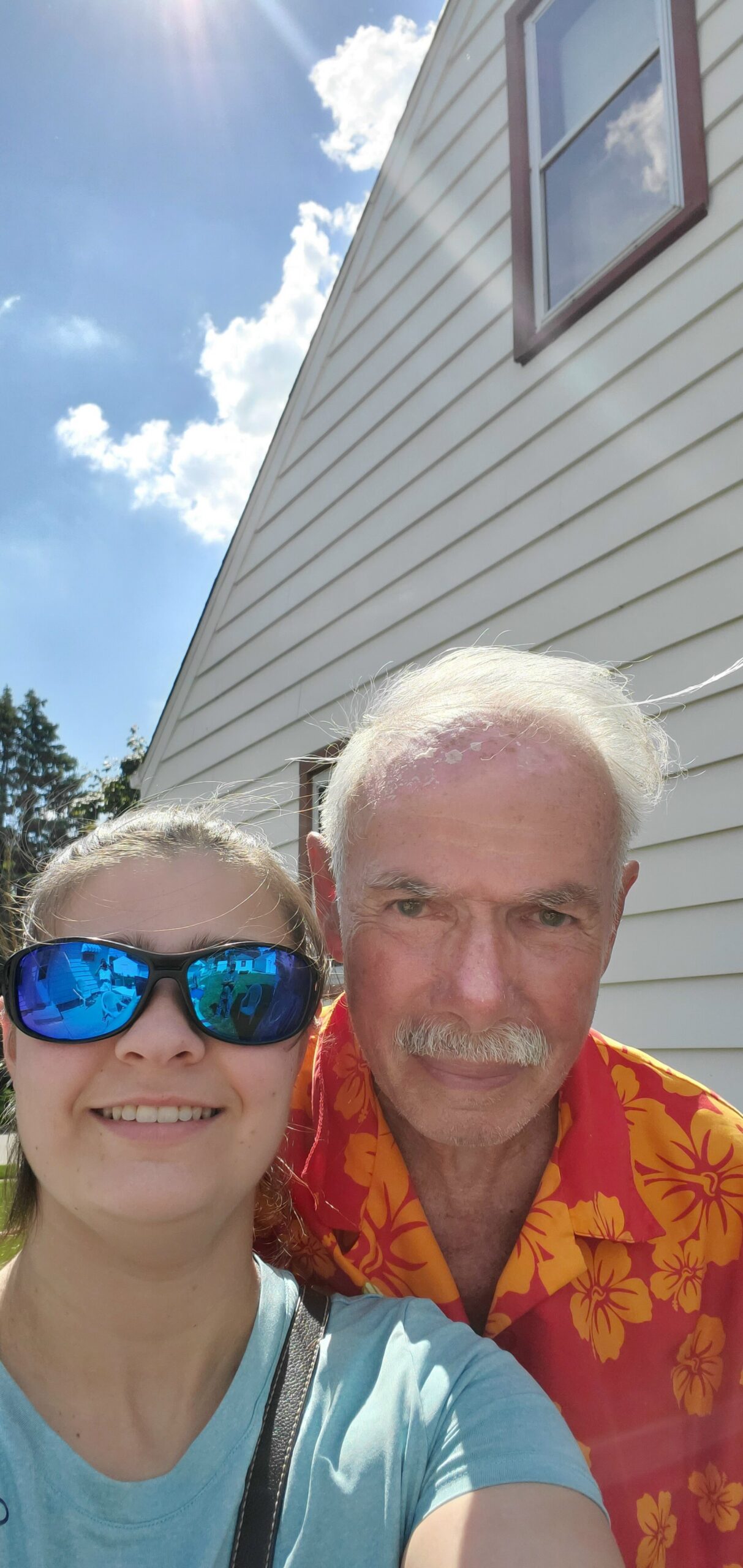 Selfie of a woman wearing blue reflective sunglasses in the foreground, with an older man in an orange hibiscus shirt behind her, outdoors by a light-sided house on a sunny day.
