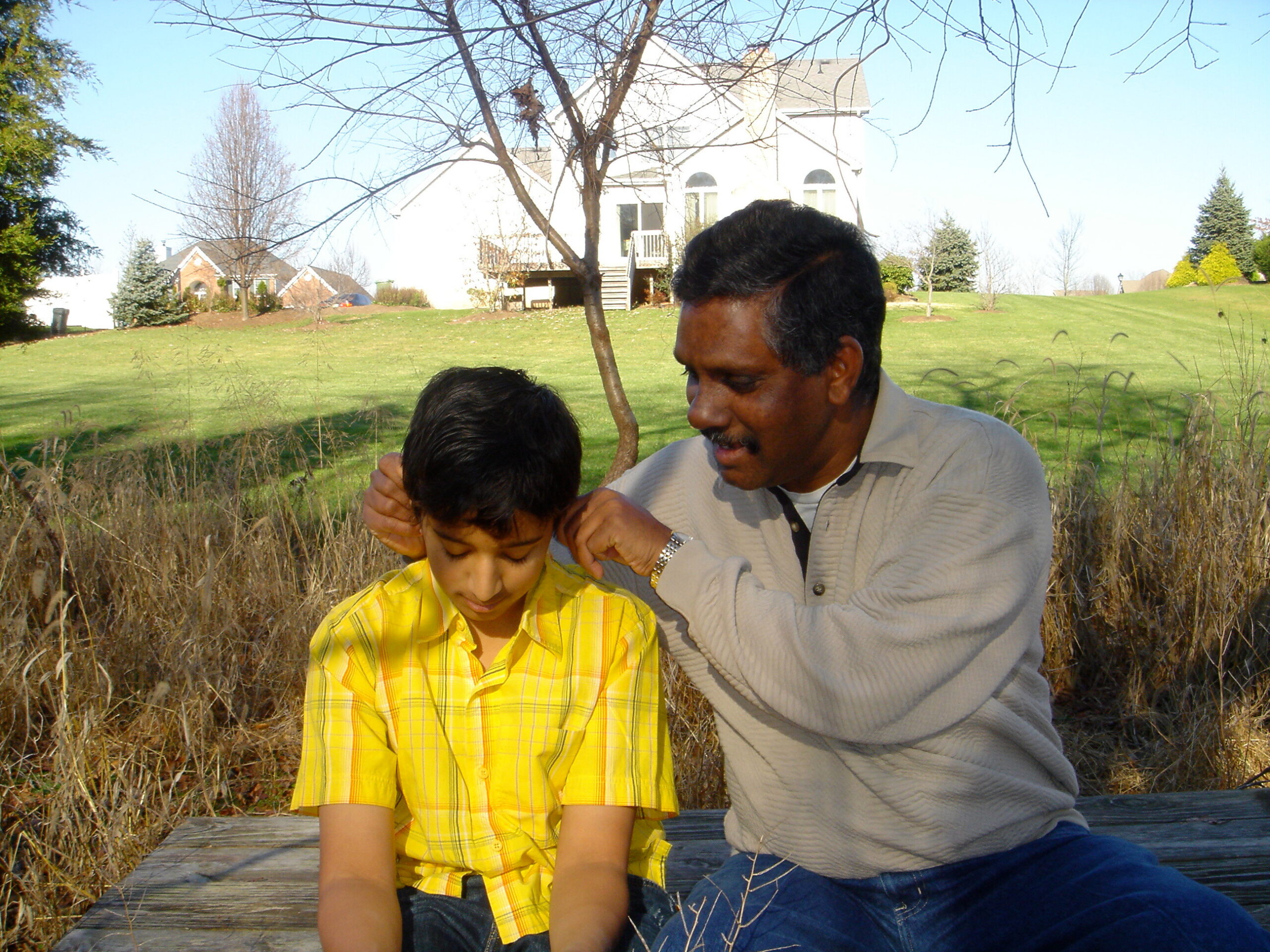 Adult man and boy sit on a wooden bench in a sunny backyard, the man adjusting the boy's hair with a white house and trees in the background.