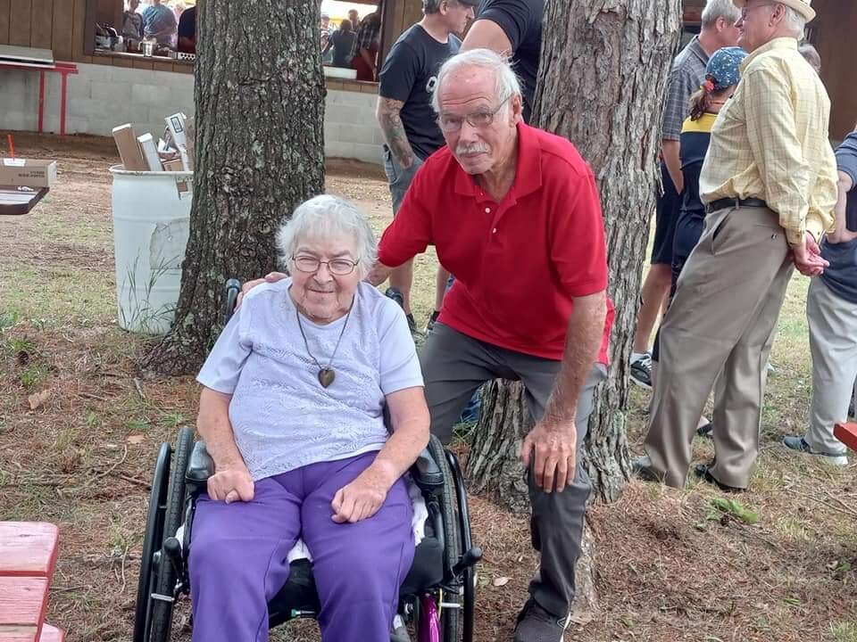 Elderly woman in a wheelchair smiles while a man in a red polo stands beside her outdoors at a park gathering.