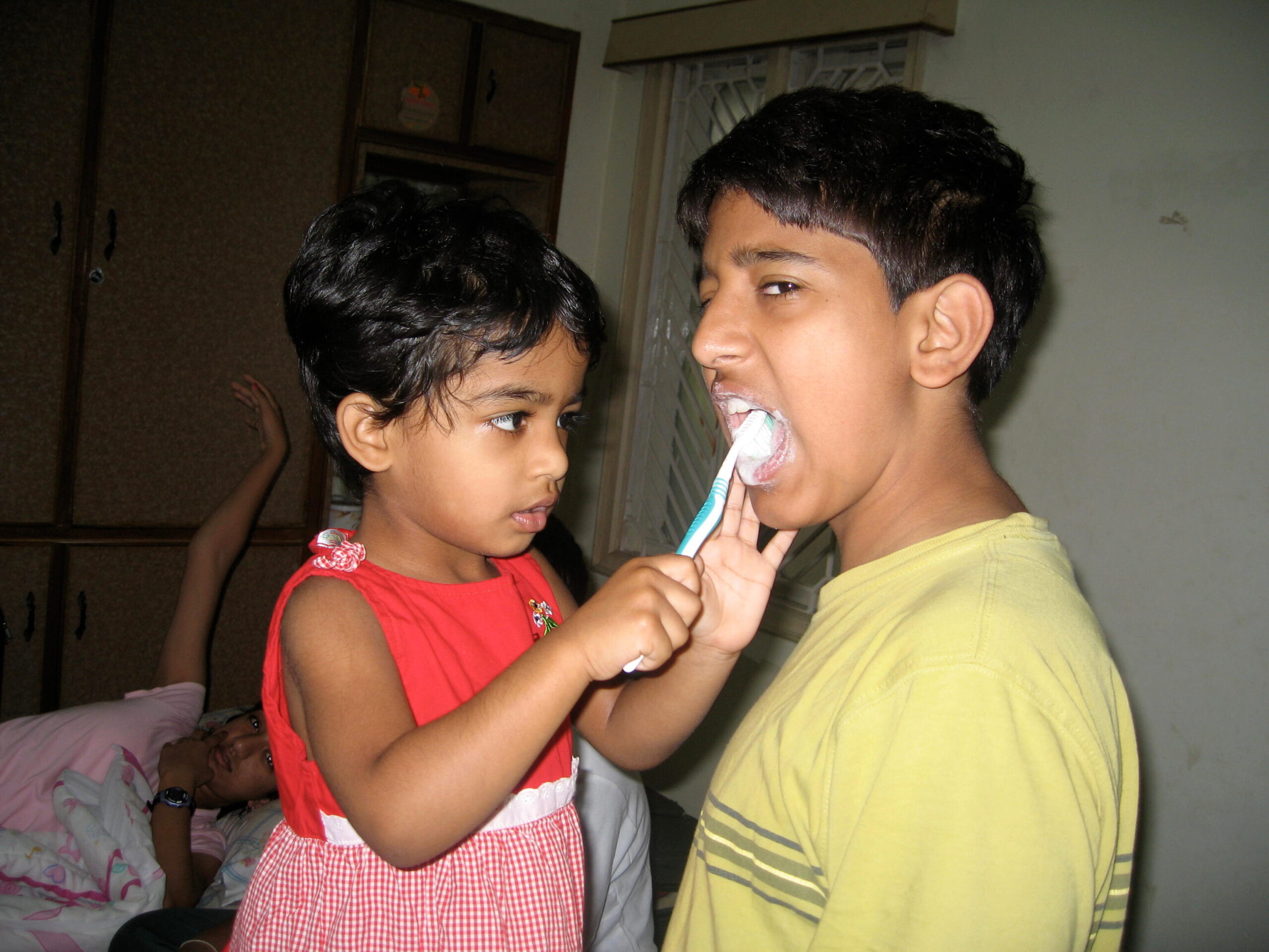 Two children brush each other's teeth indoors; girl in red dress holds a toothbrush to the boy's teeth while facing him.