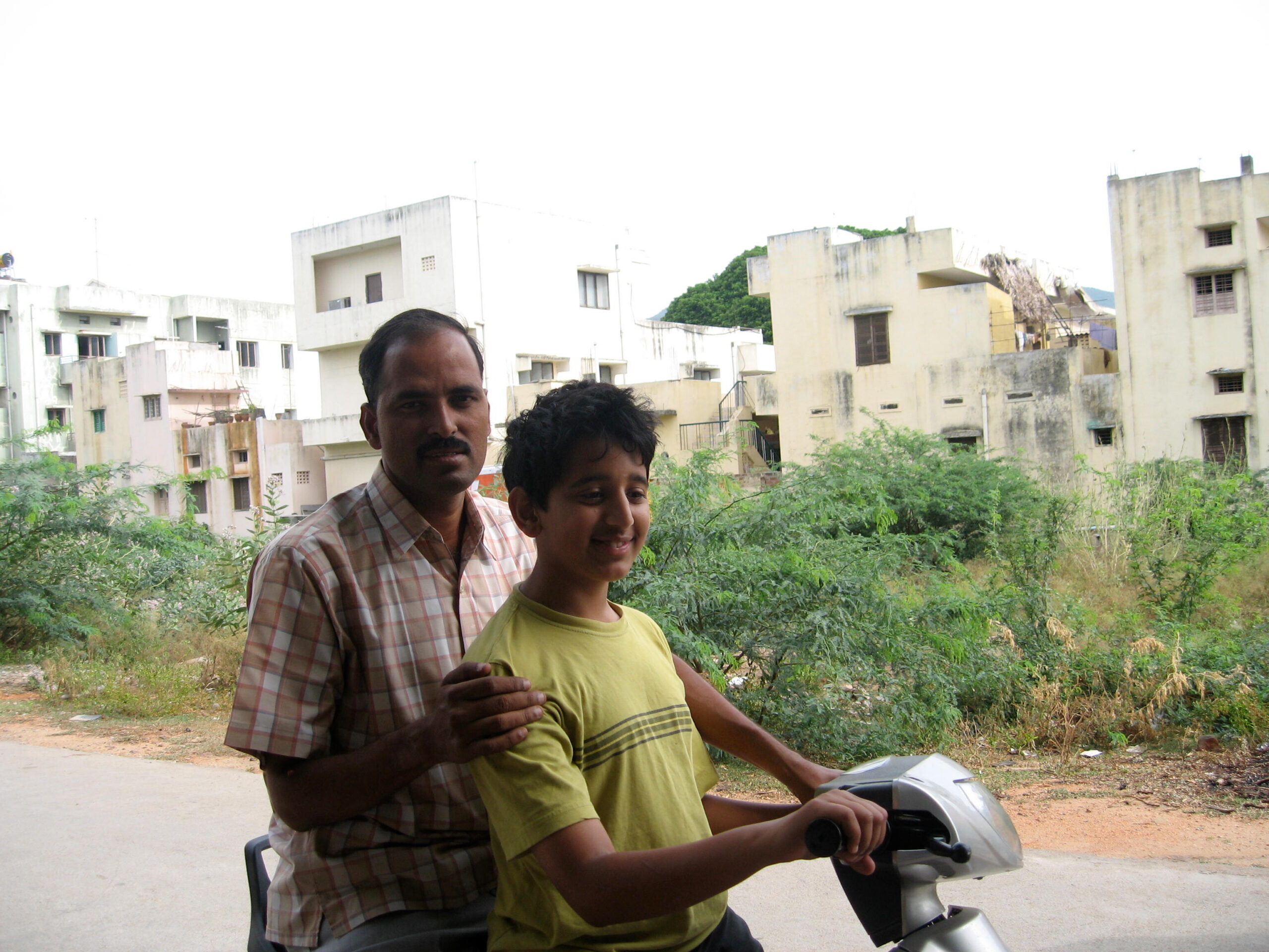 Two people riding a scooter together in a residential area with beige apartment buildings and greenery in the background.] ,