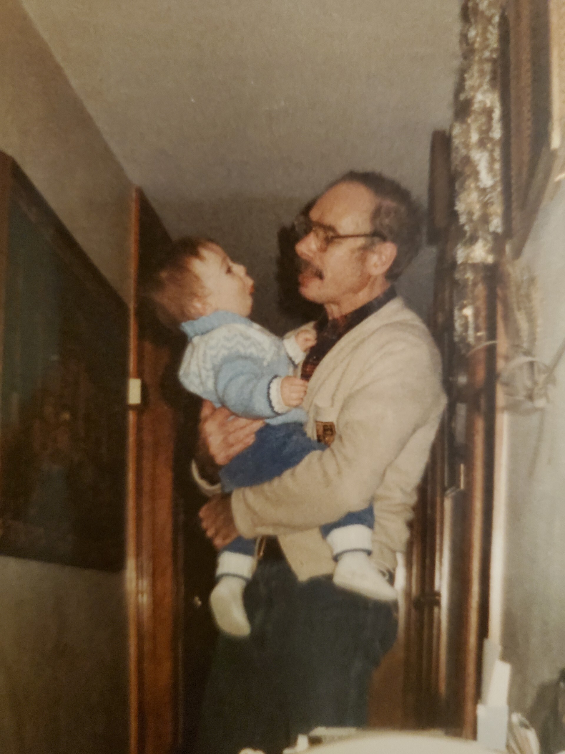 Man wearing glasses holds a smiling baby in a blue sweater indoors, facing each other lovingly.