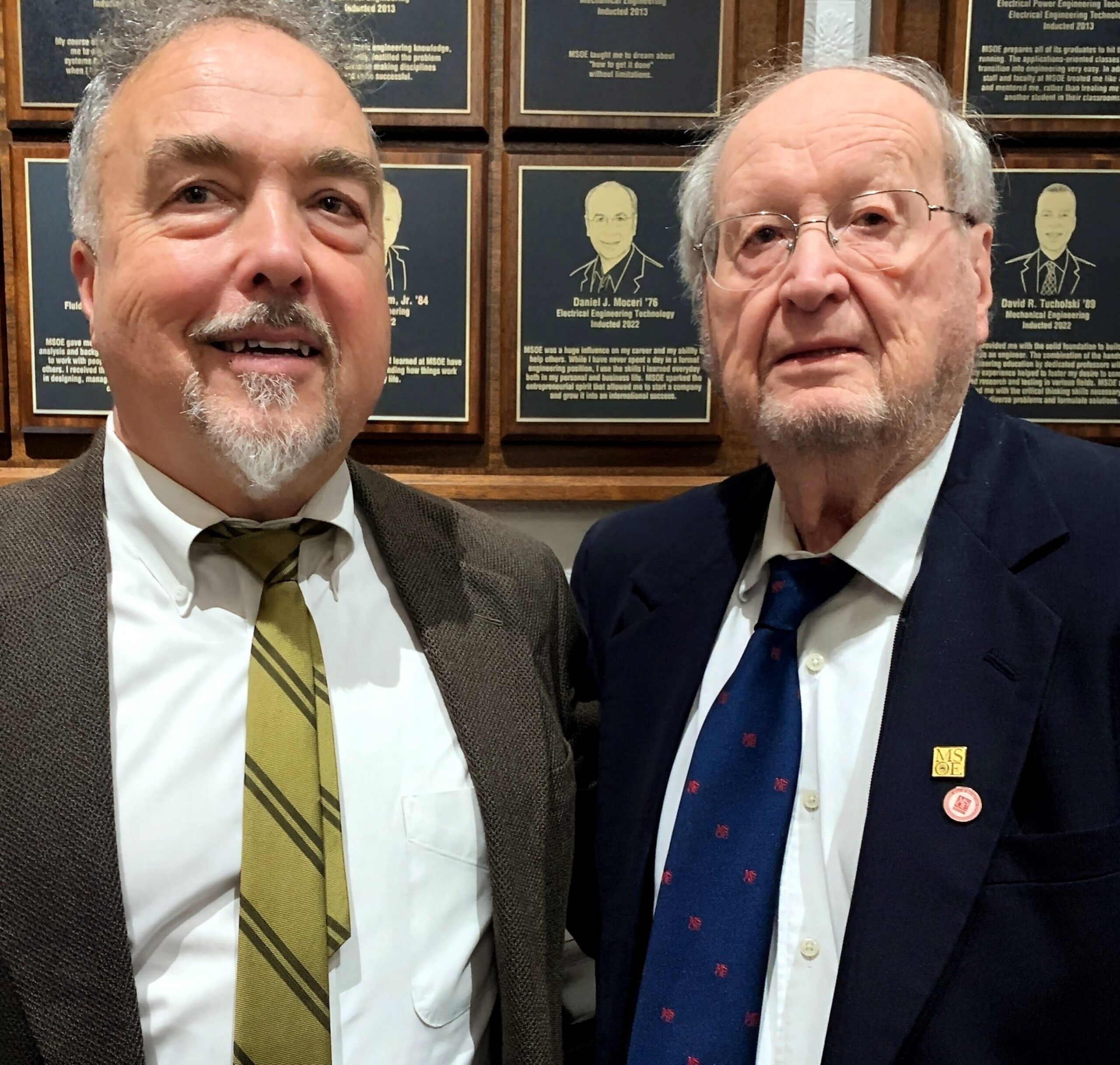 Two men in suits take a selfie in front of a wall of plaques with framed portraits behind them, smiling.