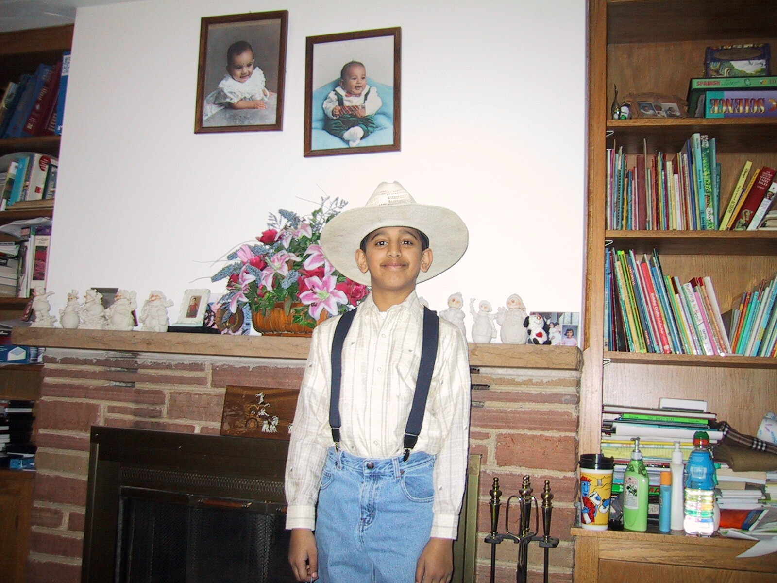 Boy wearing a white cowboy hat, striped shirt, and suspenders stands in front of a brick fireplace with a flower arrangement and family photos on the mantel.