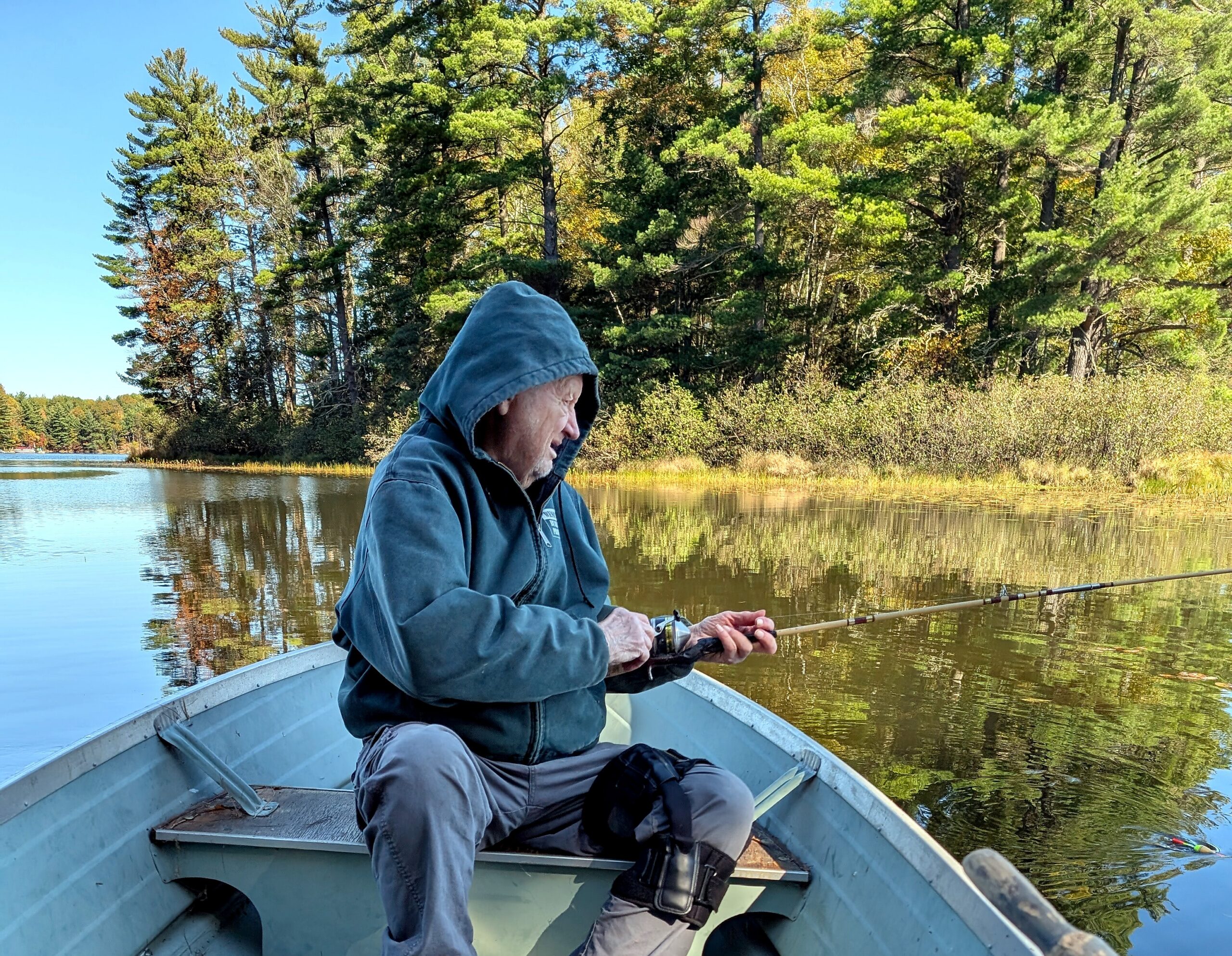 Older man in a blue hoodie fishing from a small boat on a calm lake with a tree-lined shore behind him. (informative)