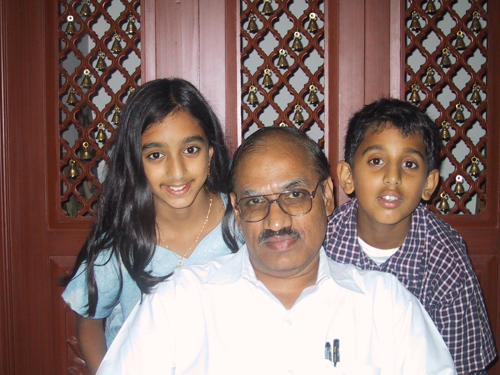 Man with glasses and mustache posing with a girl on his left and a boy on his right in front of decorative wooden panels with bells.
