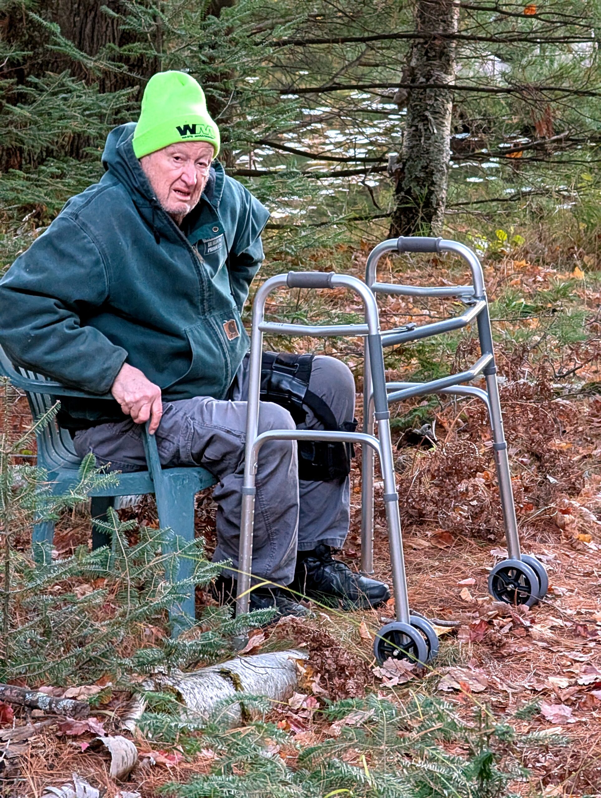 Older man in a bright green beanie sits on a green bench outside, with a metal walker nearby among pine trees and fallen leaves.