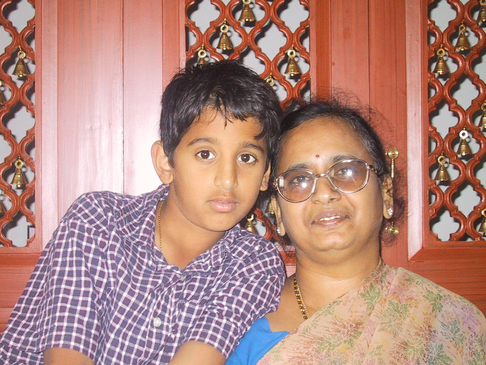 A boy and an older woman posing closely together in front of a red decorative lattice screen with bells, both looking at the camera.
