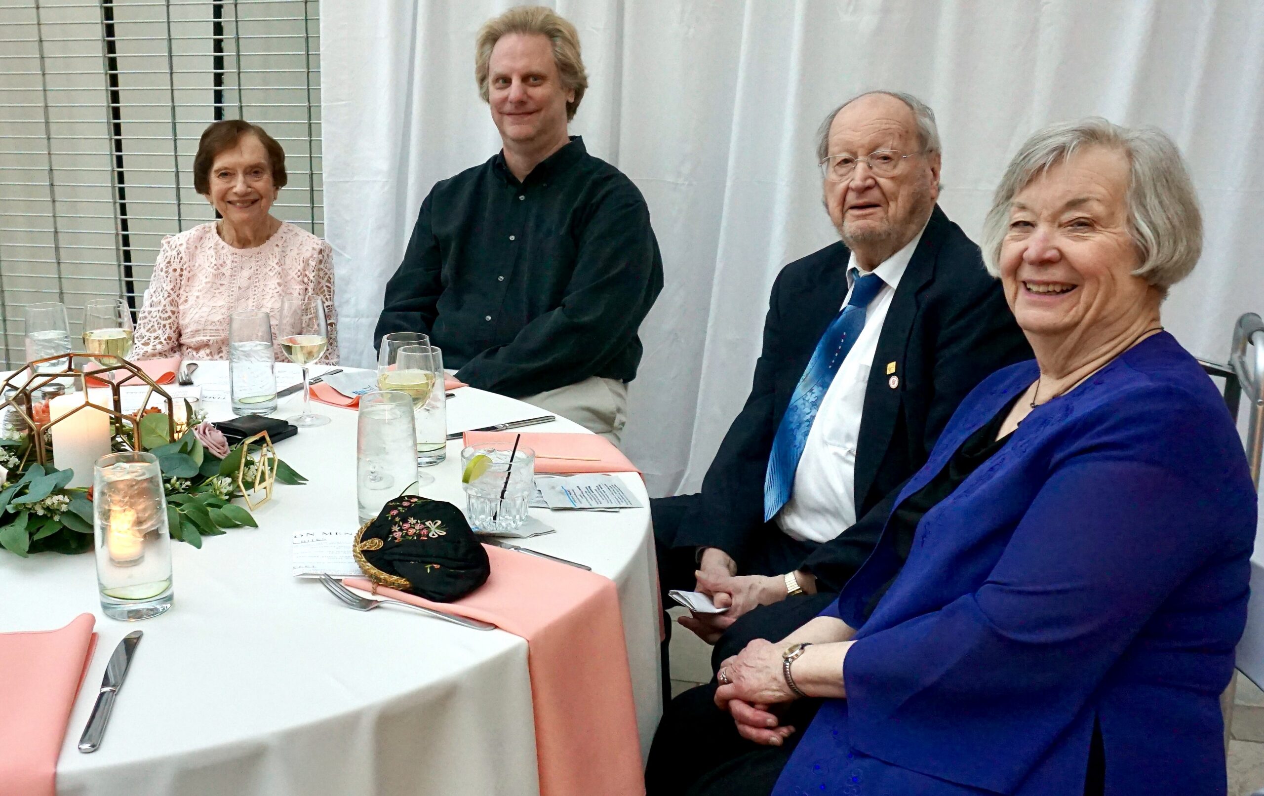 Four adults sit at a round dining table, smiling for the camera; a woman in a pink lace top is on the left, a man in a black shirt sits next to her, an older man in a suit and blue tie, and a smiling woman in a blue cardigan on the right; table is set with pink napkins, glassware, and a floral centerpiece.