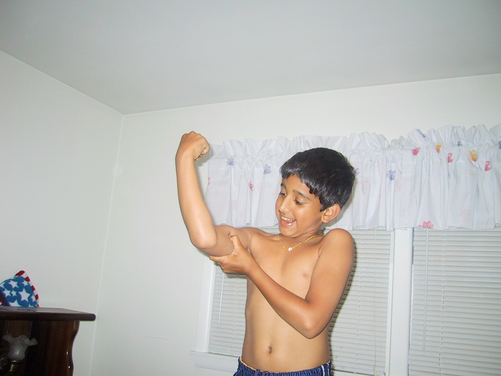 Boy indoors, shirtless, smiling while flexing his arm at shoulder height near a window with blinds and a ruffled valance in the background.