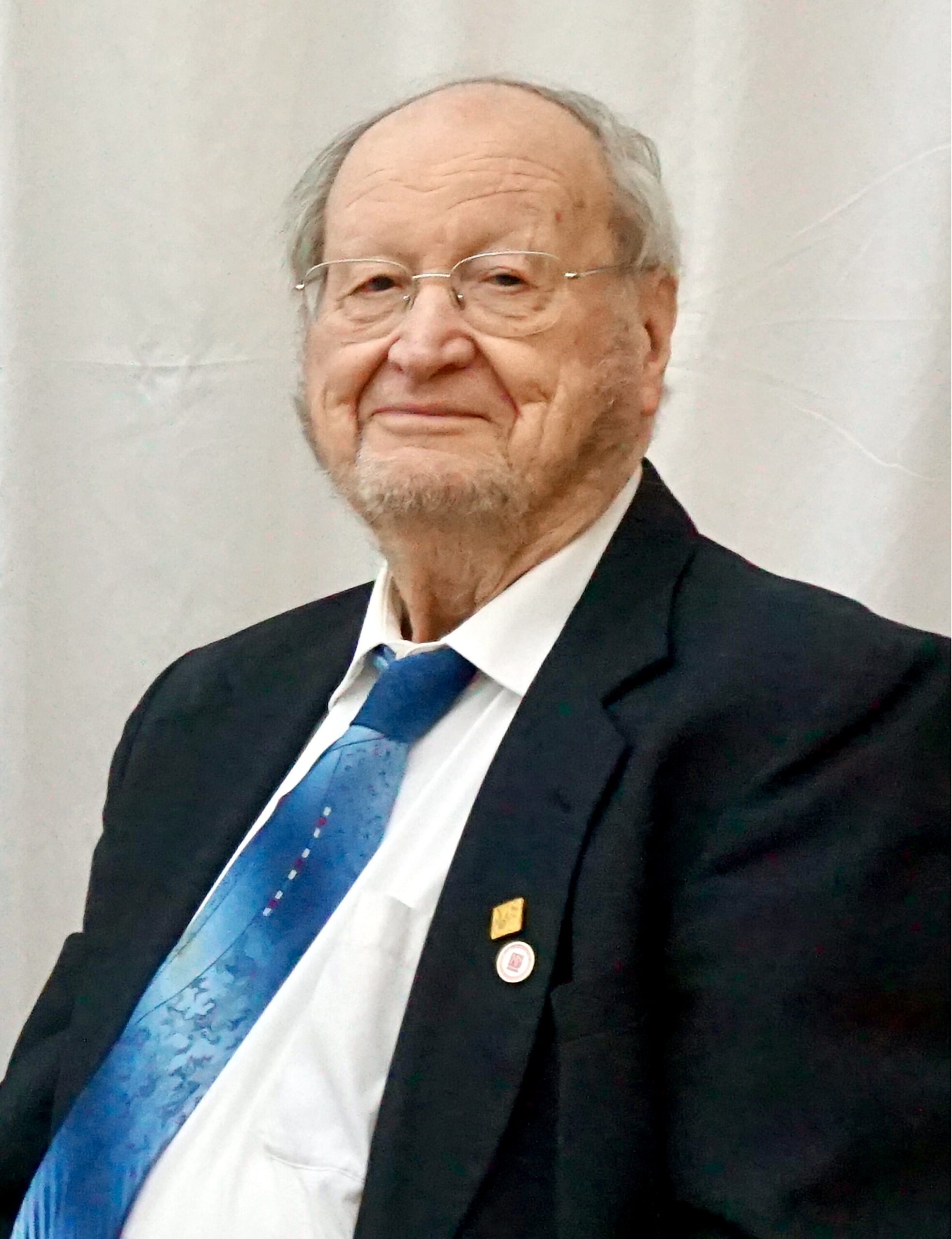 Older man in a dark suit and blue patterned tie, smiling at the camera, wearing glasses and lapel pins.