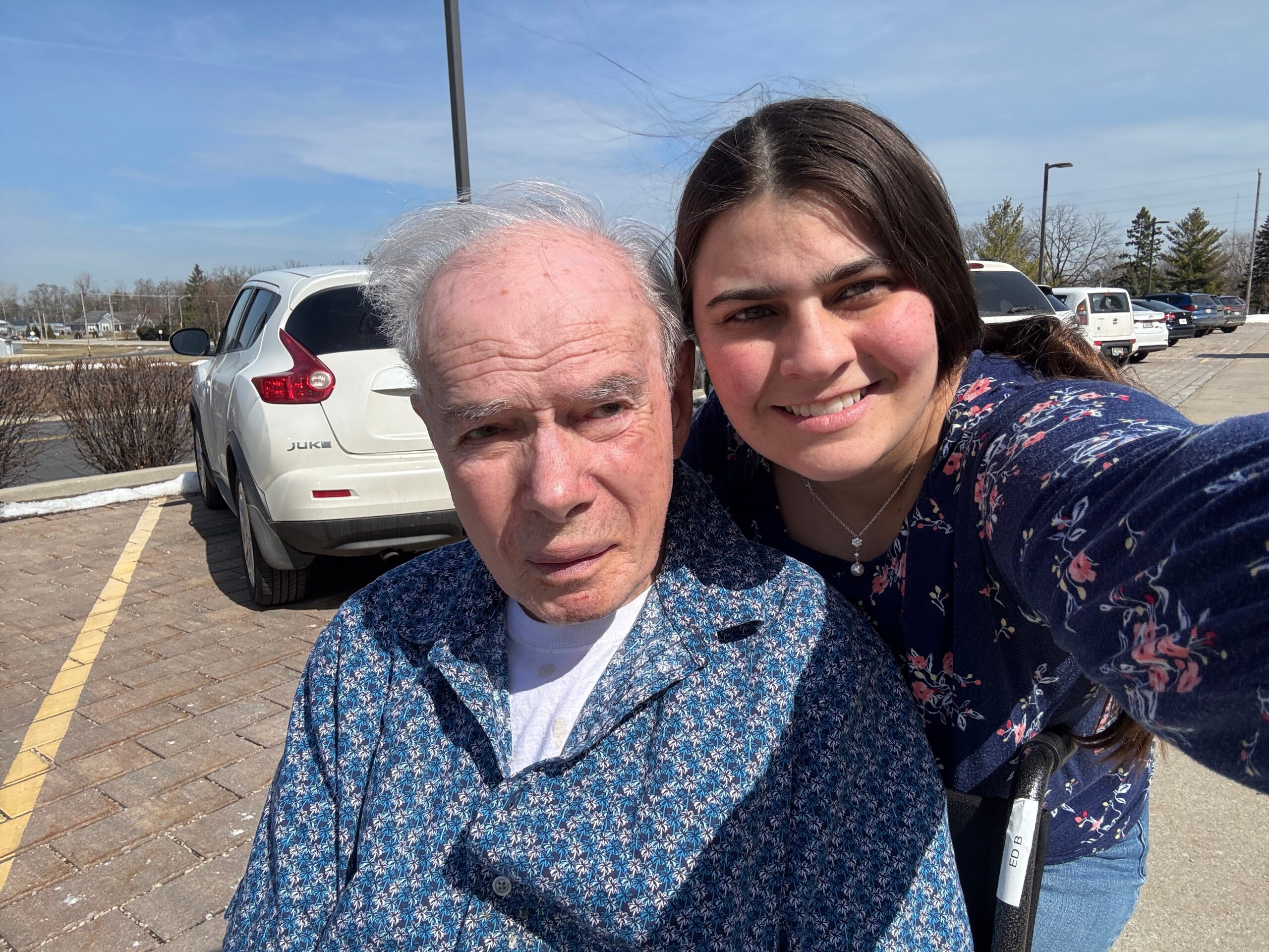 Two people take a sunny outdoor selfie in a parking lot: an older man in a blue patterned shirt and a smiling younger woman beside him.