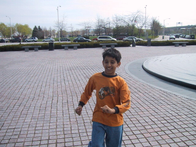 Smiling boy in an orange shirt and jeans stands on a paved plaza beside a circular fountain.