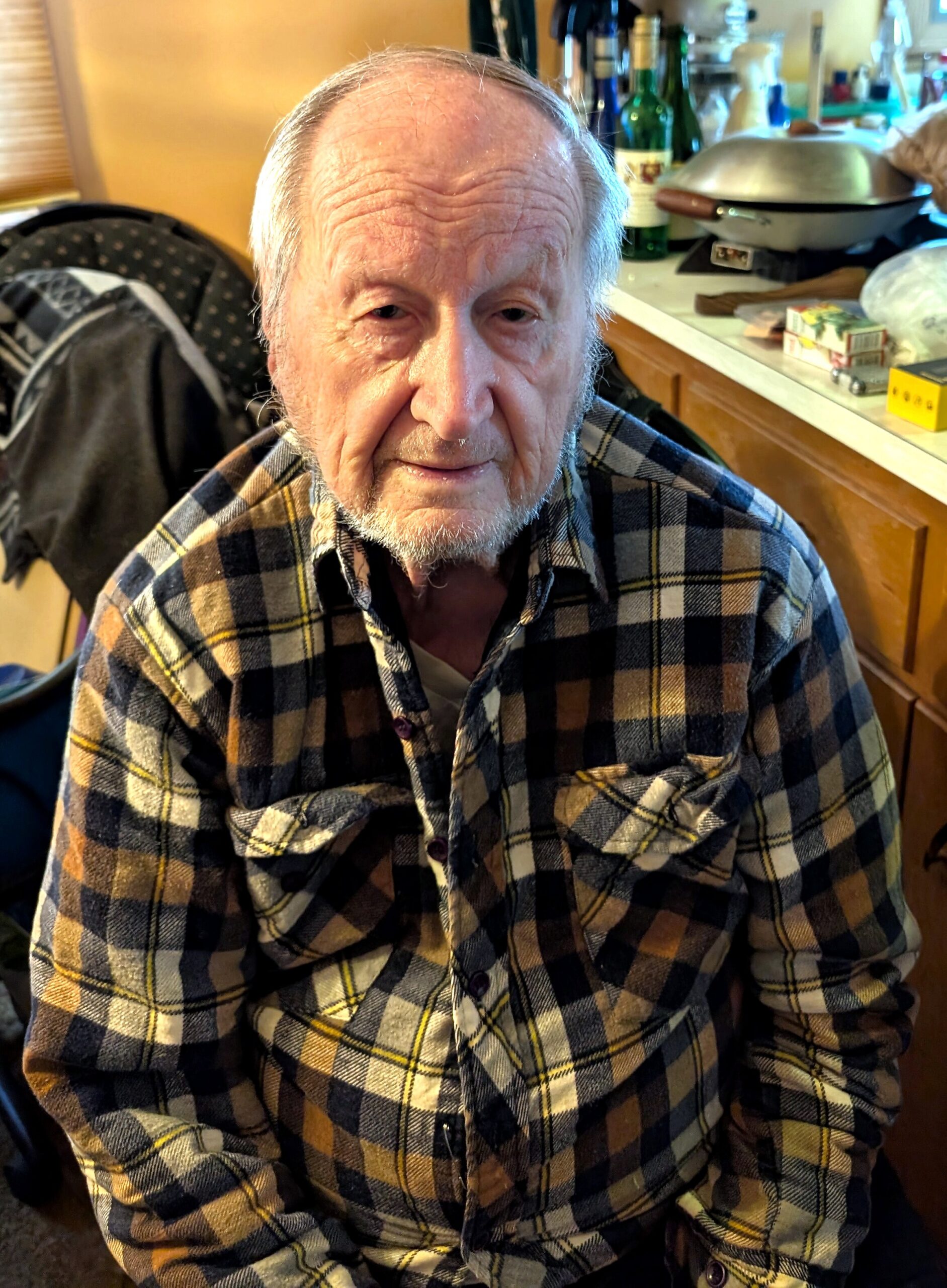 Close-up of an elderly man with a gray beard wearing a blue plaid shirt, seated in a kitchen.