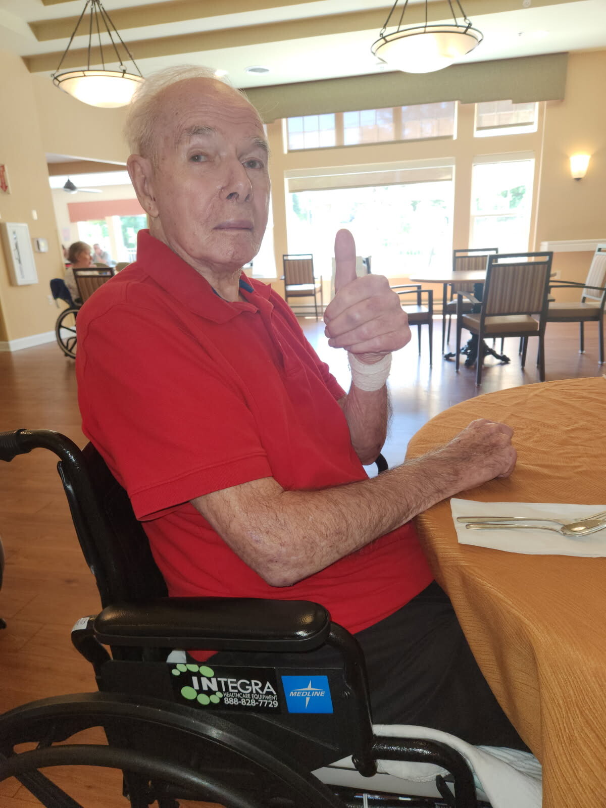 Elder man in a red polo shirt sits in a wheelchair at a dining table, giving a thumbs-up with his bandaged right hand in a bright, shared dining room.