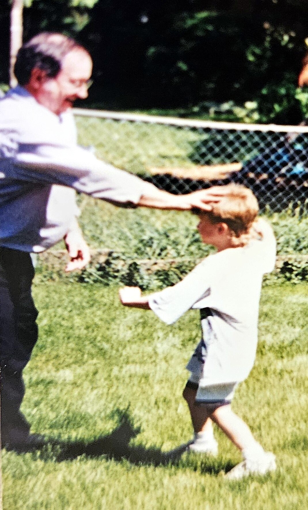 Older man in a light shirt pats a young boy on the head in a sunlit grassy yard, with a chain-link fence in the background.