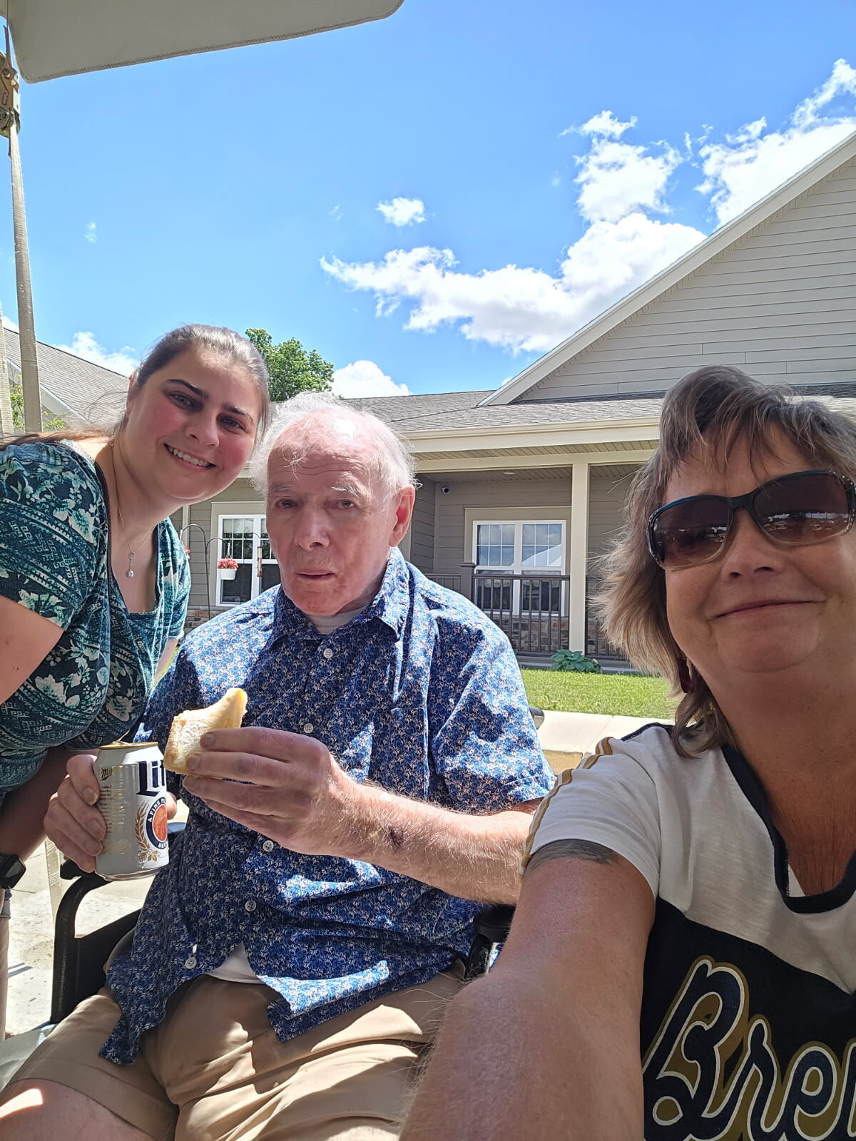Three people smiling in a sunny backyard; an older man in a blue floral shirt holds a piece of bread and a can, with two women on either side.