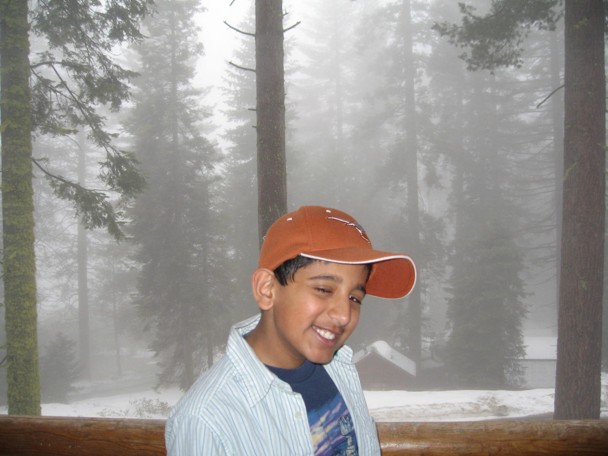 Smiling boy in an orange cap and striped shirt outdoors in a snowy, foggy forest.