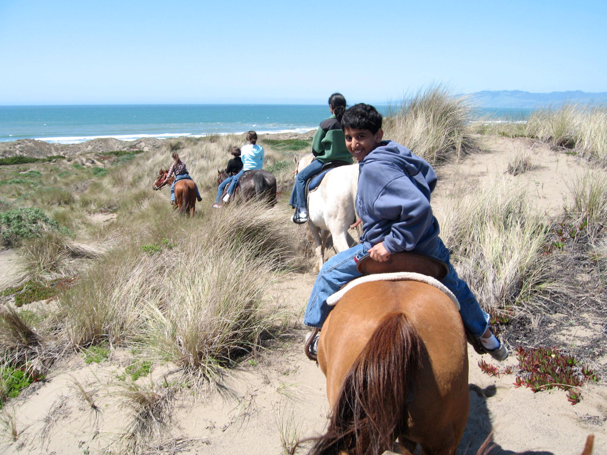 People horseback riding along a sandy beach with grasses, the ocean visible in the distance and a smiling rider in the foreground.