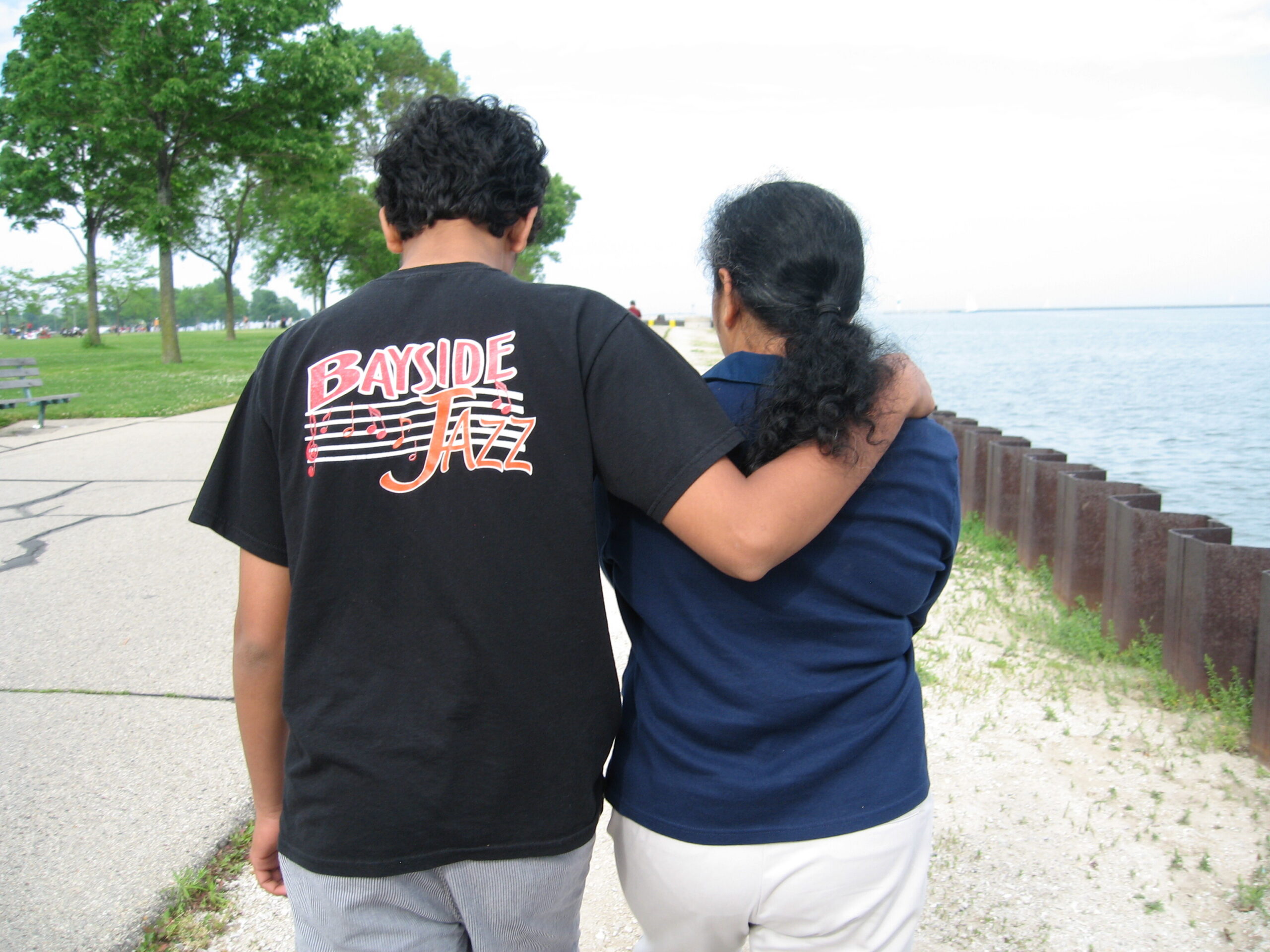 Two people walk along a lakeside path, seen from behind; one wearing a black T-shirt with 'BAYSIDE JAZZ' and the other in a blue top, arms around each other.