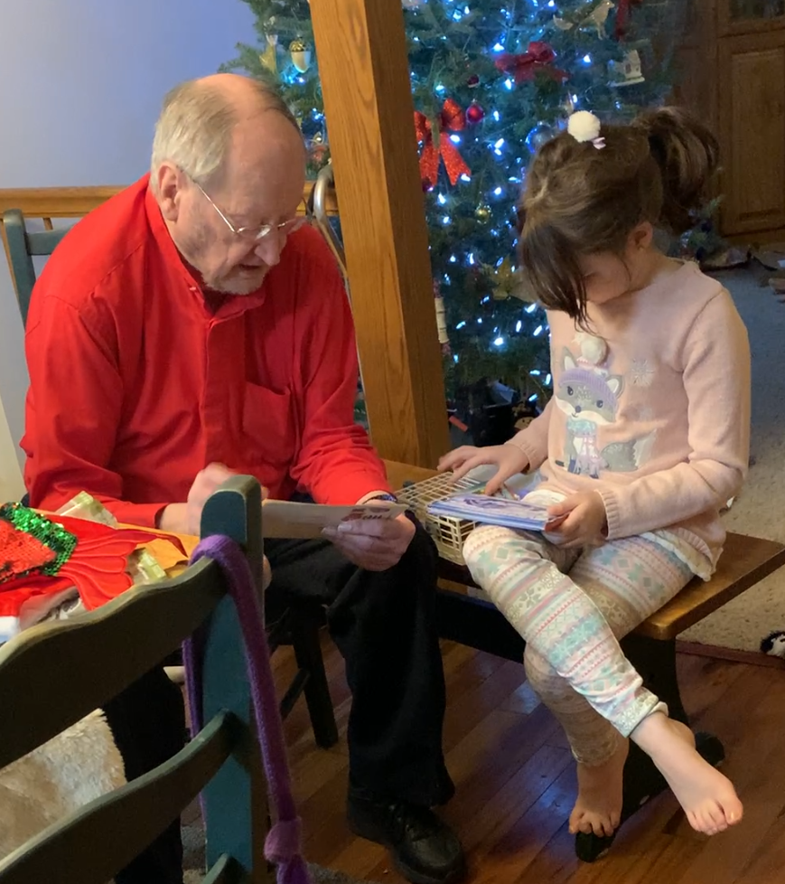 Grandfather and young girl reading a book together beside a lit Christmas tree.