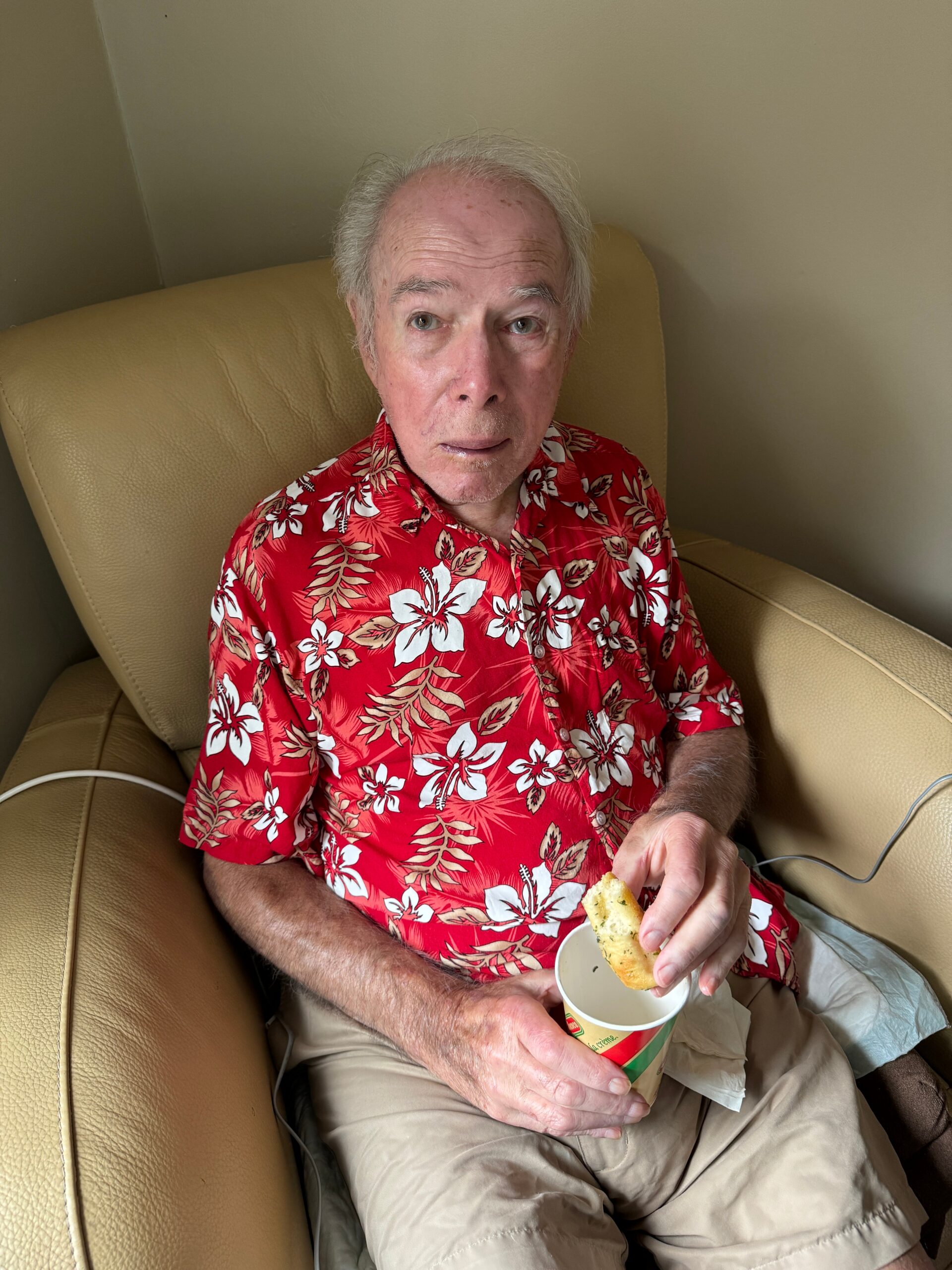 Elderly man with white hair sits on a beige leather chair, wearing a red tropical shirt, holding a cup and a piece of bread/snack.