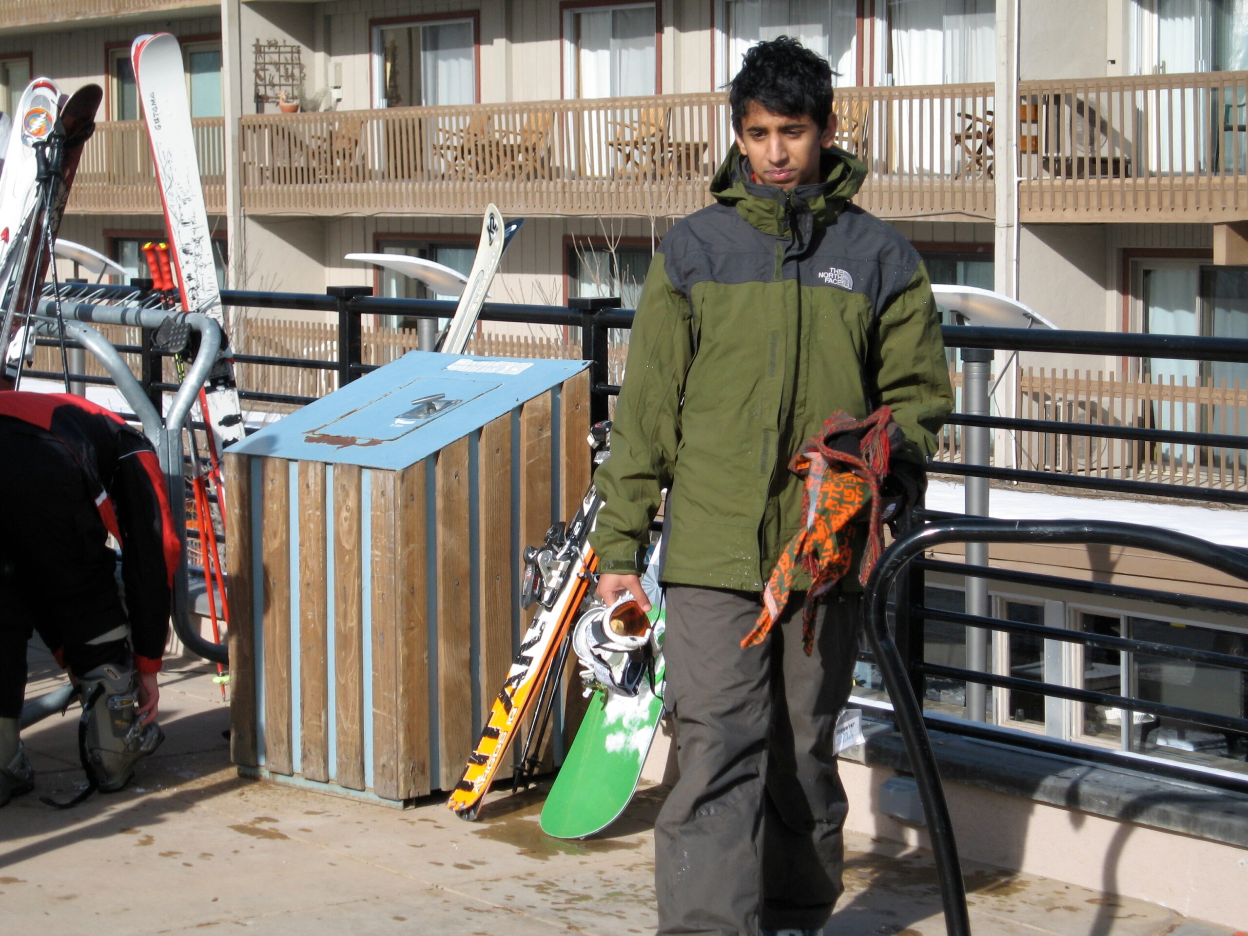 Young man in a green and gray winter jacket stands with a green snowboard at a ski rack outdoors, holding gloves in one hand.
