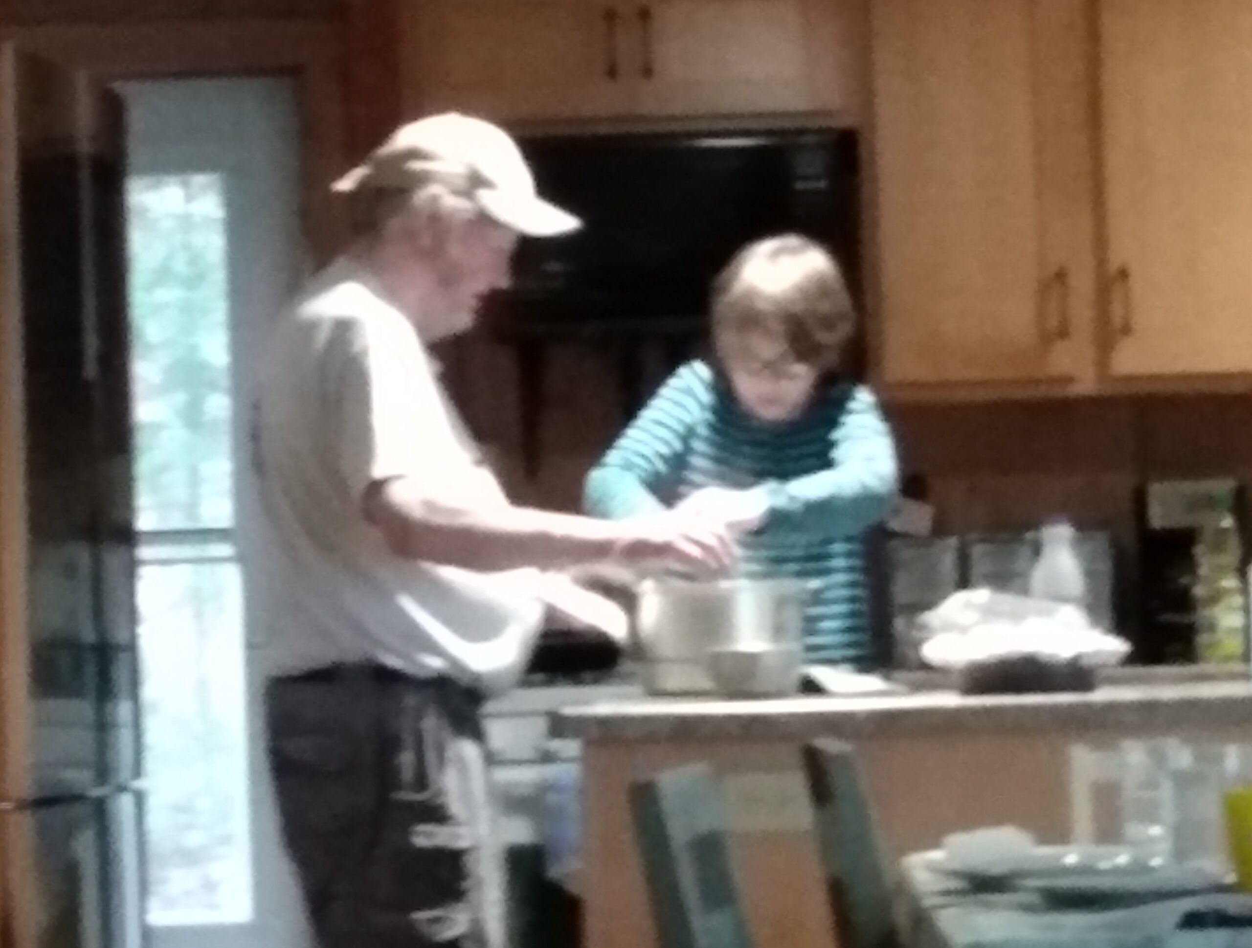 Two people cooking together in a home kitchen, one in a light shirt and cap, the other in a striped shirt, near a counter with dishes.