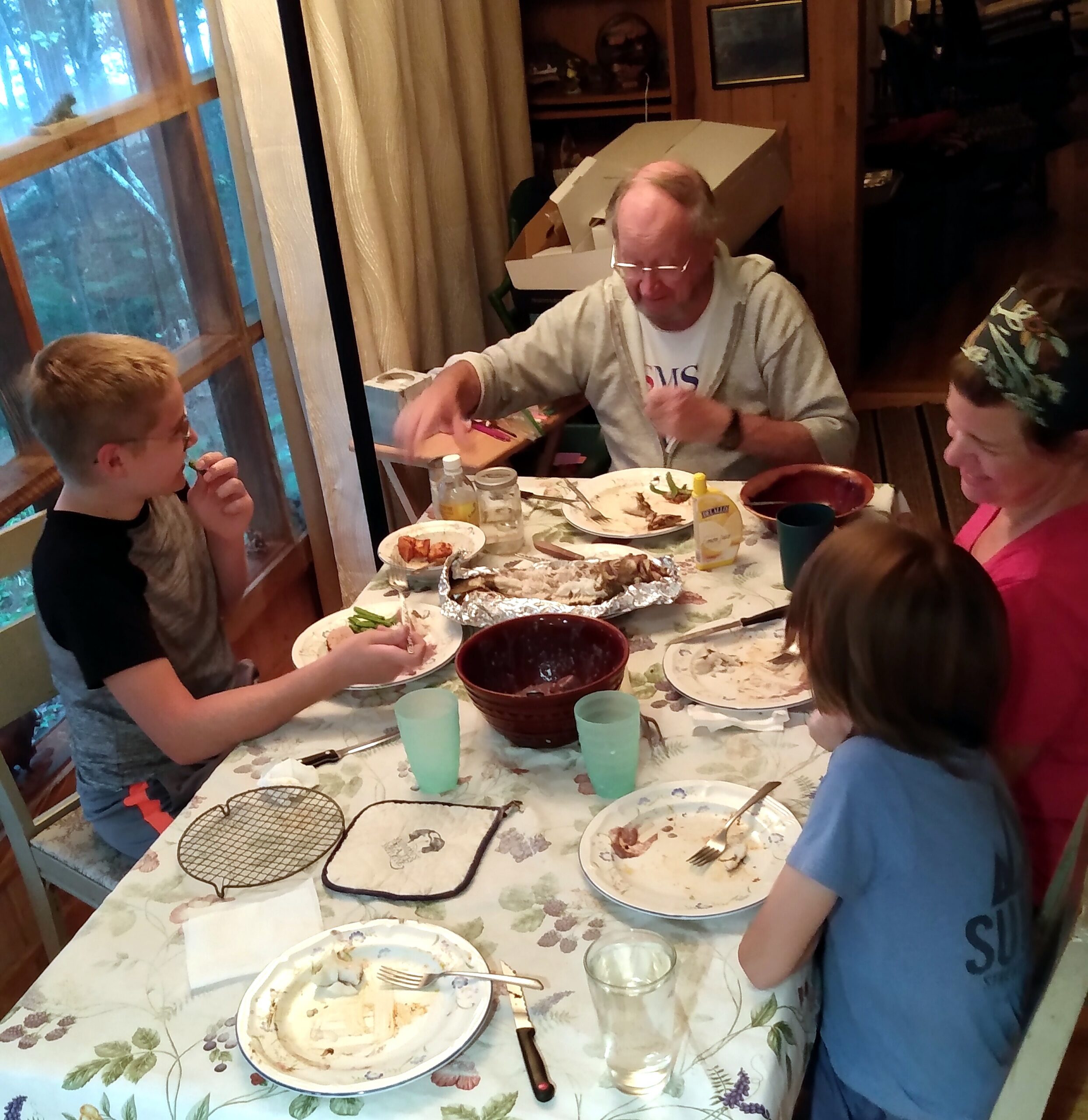 People seated around a dining table eating, with plates, cups, and foil-wrapped dishes visible in a home setting.