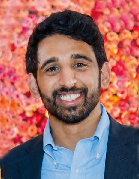 Smiling man with dark hair and beard, wearing a light blue shirt and dark blazer, in front of a pink flower backdrop
